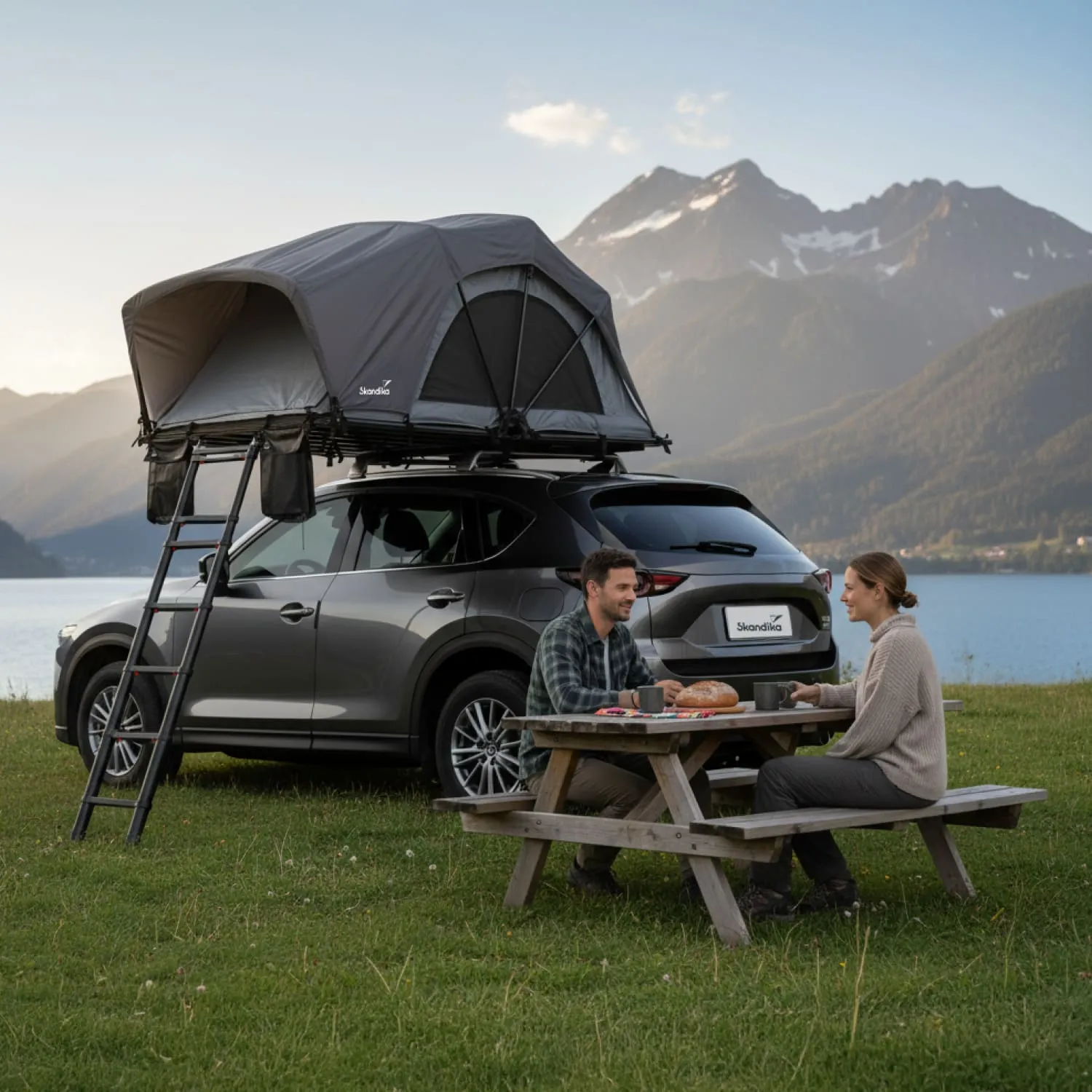 Zwei Personen sitzen an einem Holztisch mit Essen und Getränken. Im Hintergrund steht ein Auto mit einem Dachzelt, vor einer Berglandschaft.