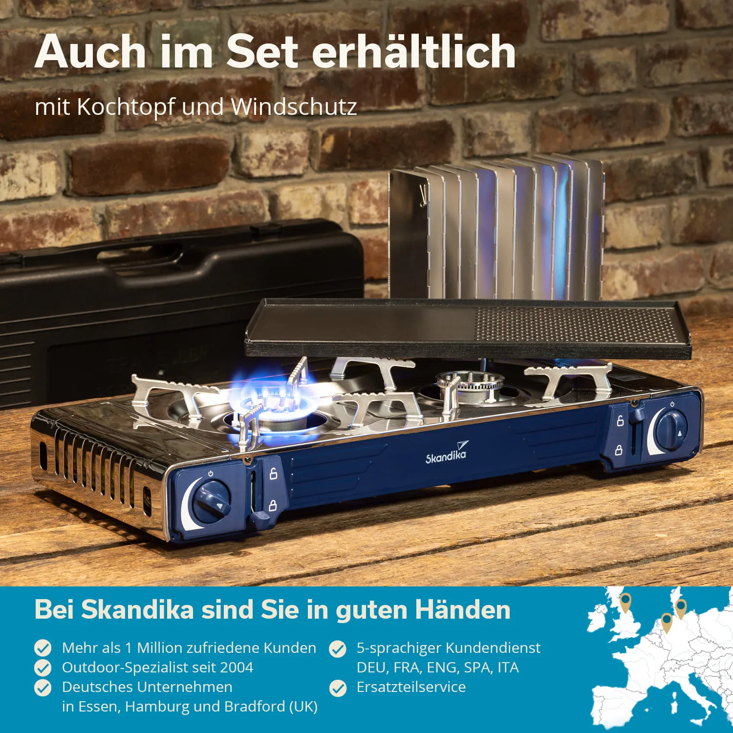 Campingkocher mit Flamme und Windschutz auf einem Holztisch, im Hintergrund eine gemauerte Wand. Zubehör sichtbar.