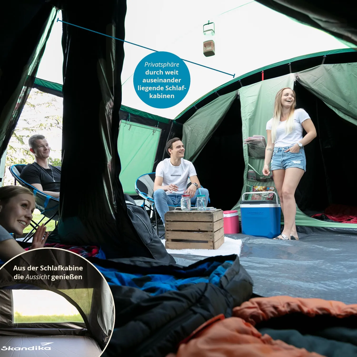 Innenansicht eines Zeltes mit mehreren Schlafkabinen. Zwei Personen sitzen, eine Person steht. Ein Tisch und Kühlbox sind sichtbar.