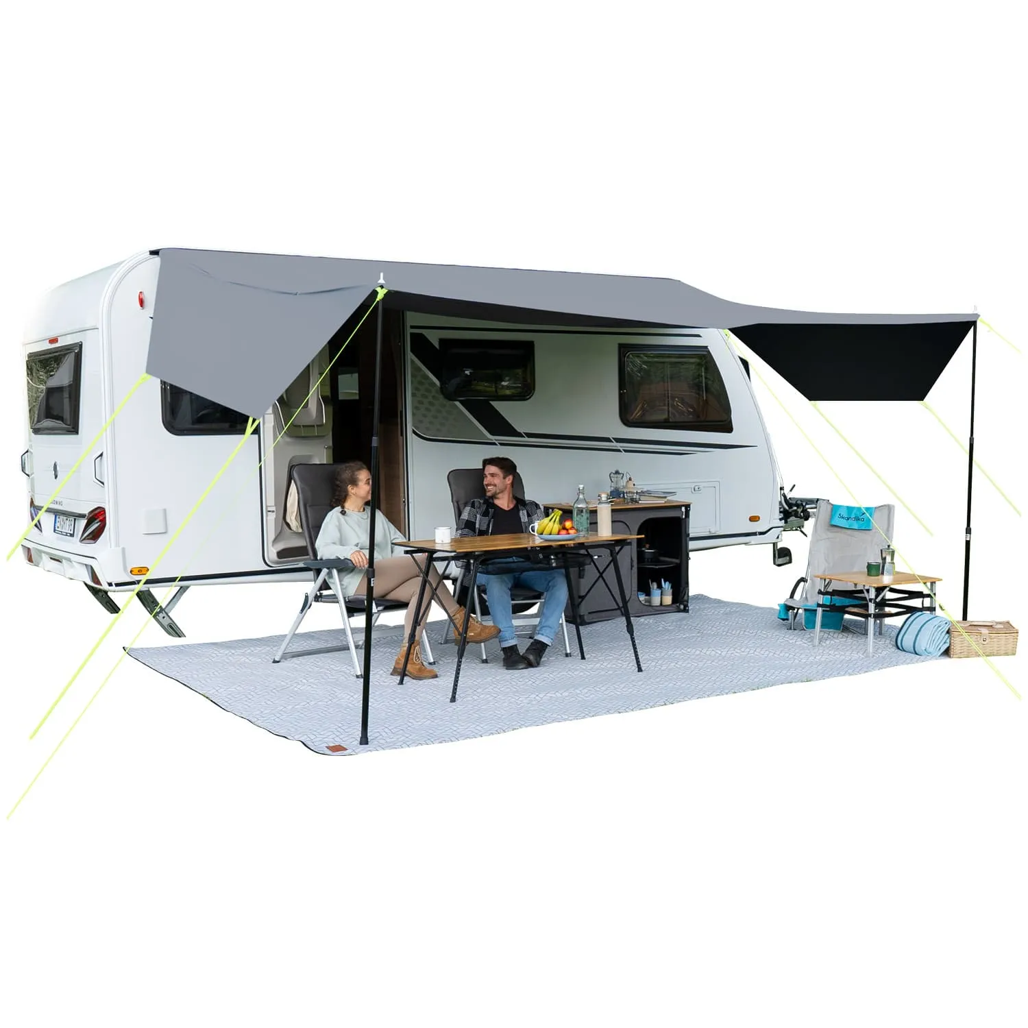 Ein Wohnwagen mit Markise. Zwei Personen sitzen an einem Tisch, umgeben von Stühlen und einem Teppich. Auf dem Tisch sind Obst und Getränke.