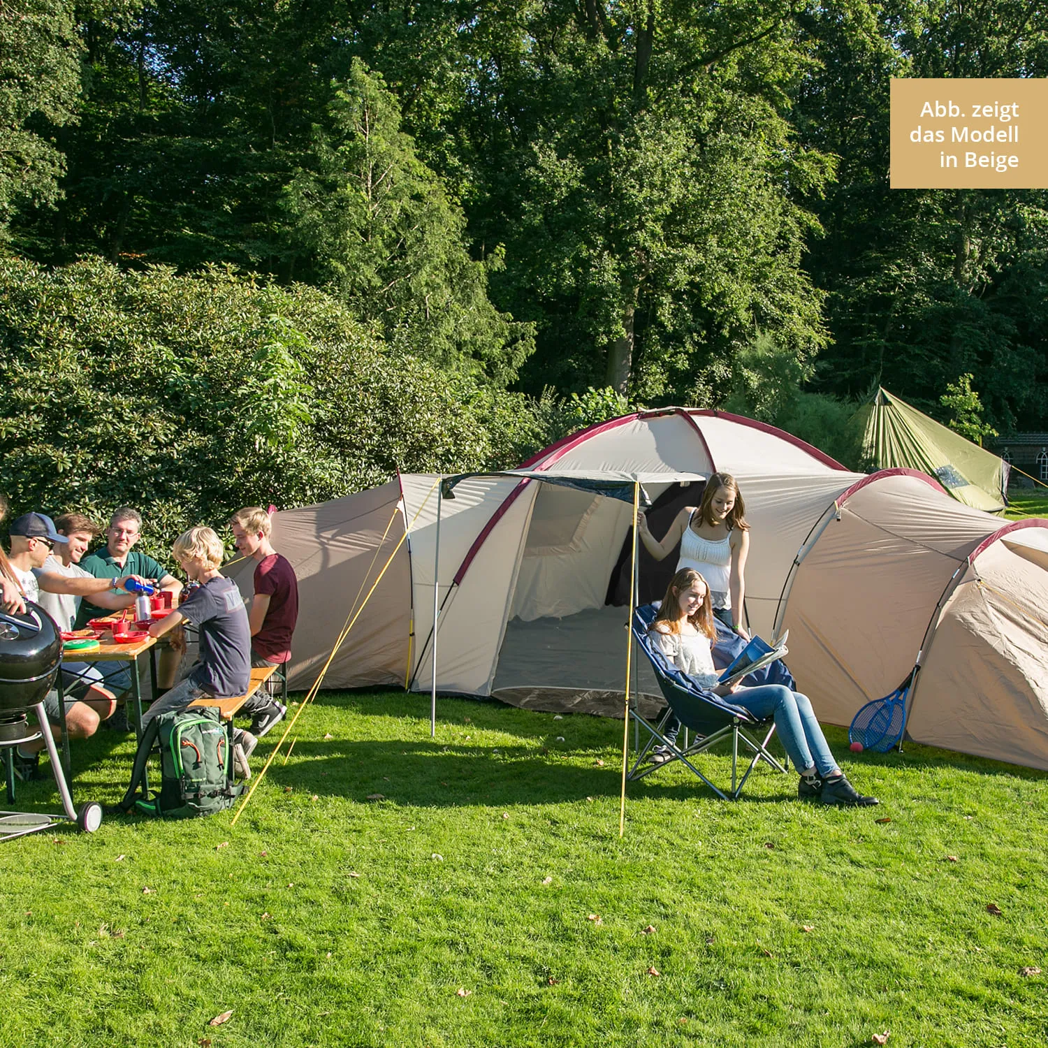 Zelt in der Natur mit mehreren Personen am Tisch, eine sitzende Person liest. Grüne Wiese, Bäume im Hintergrund.