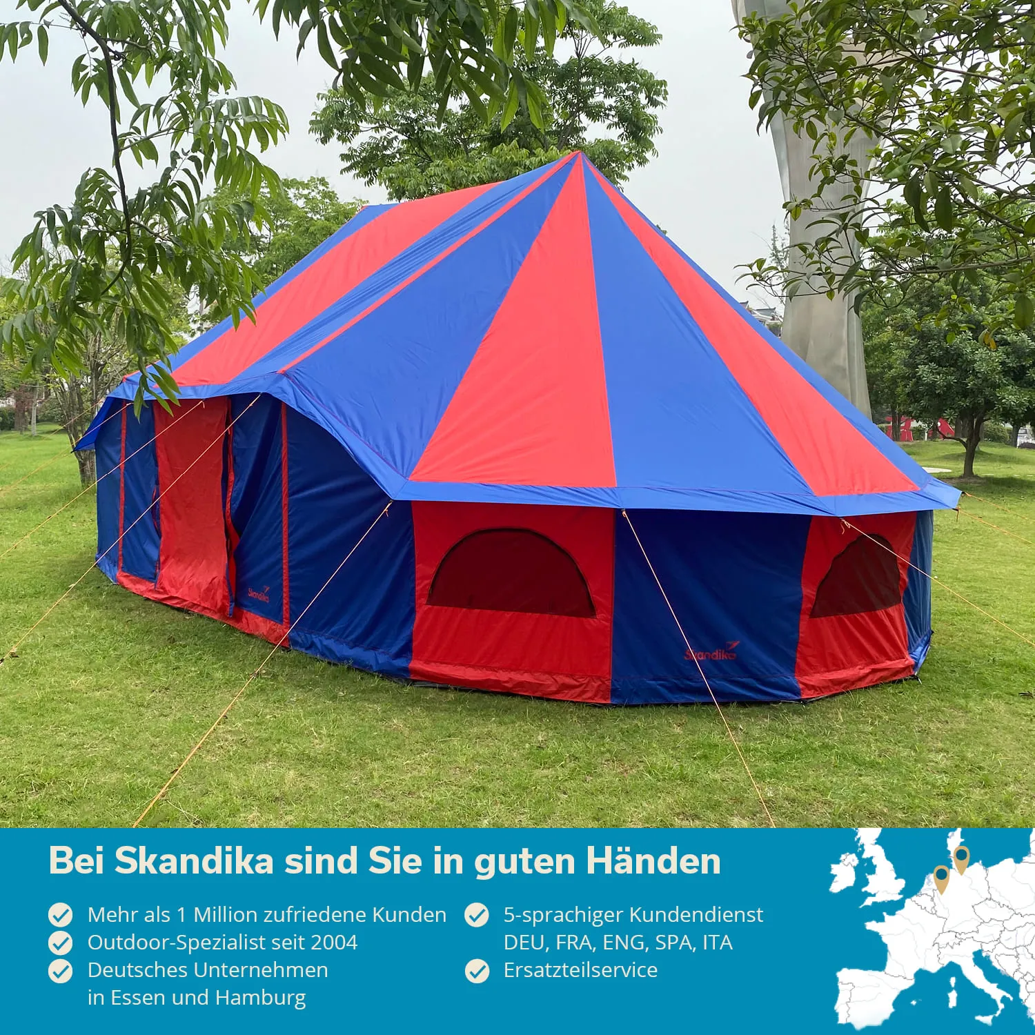 Ein großes, buntes Zelt mit blauen und roten Segmenten steht auf einer grünen Wiese, umgeben von Bäumen.