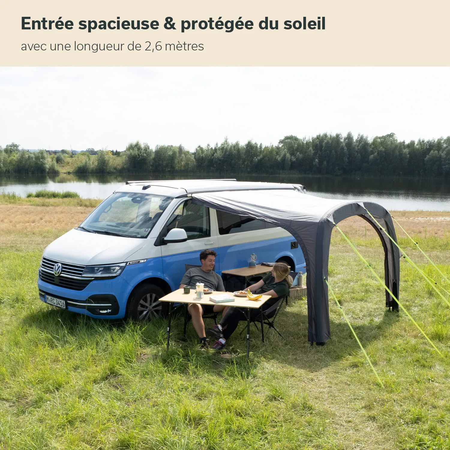 Ein blauer VW-Bus steht auf einer Wiese. Darunter ein Markisenbereich mit einem Tisch und zwei Stühlen.