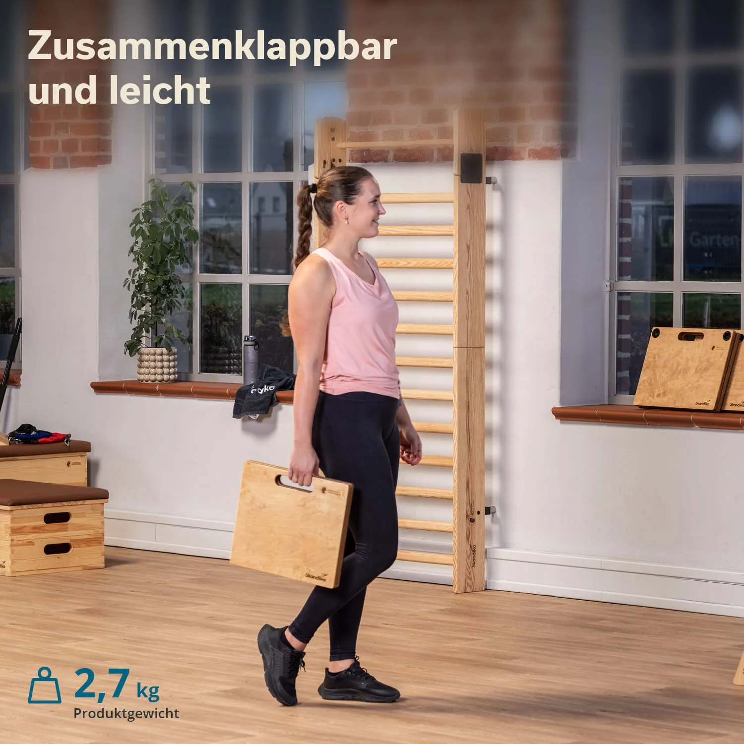 Person hält ein zusammenklappbares, leichtes Holzbrett in einem Raum mit Fenstern und Fitnessgeräten.