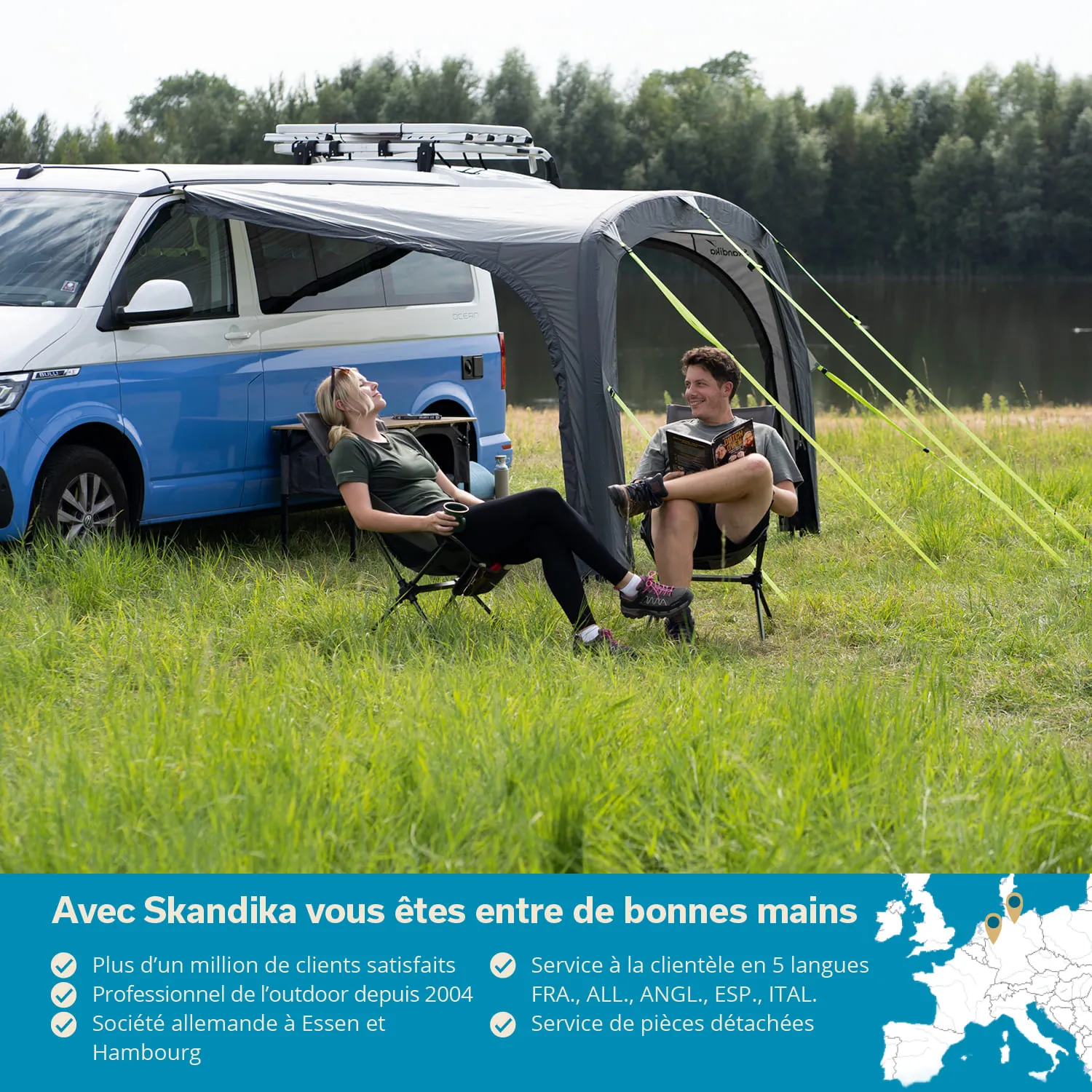 Ein blau-weißer Camper steht auf einer Wiese. Zwei Personen sitzen unter einem grauen Vorzelt, eine hat eine Tasse, die andere liest. Im Hintergrund ist ein Gewässer und Bäume zu sehen.