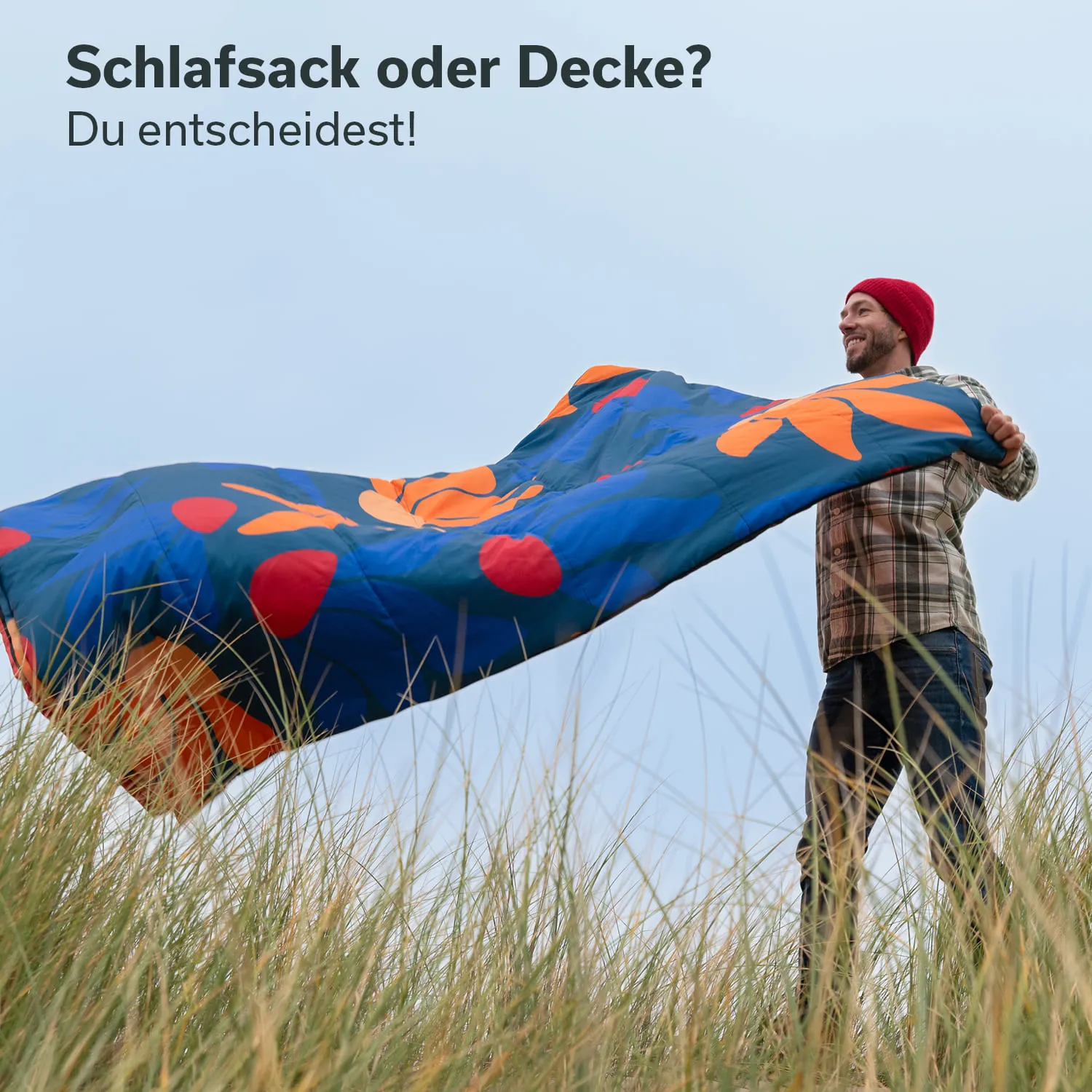 Person in kariertem Hemd und roter Mütze hält buntes Tuch in der Hand, umgeben von hohem Gras. Himmel ist bewölkt.