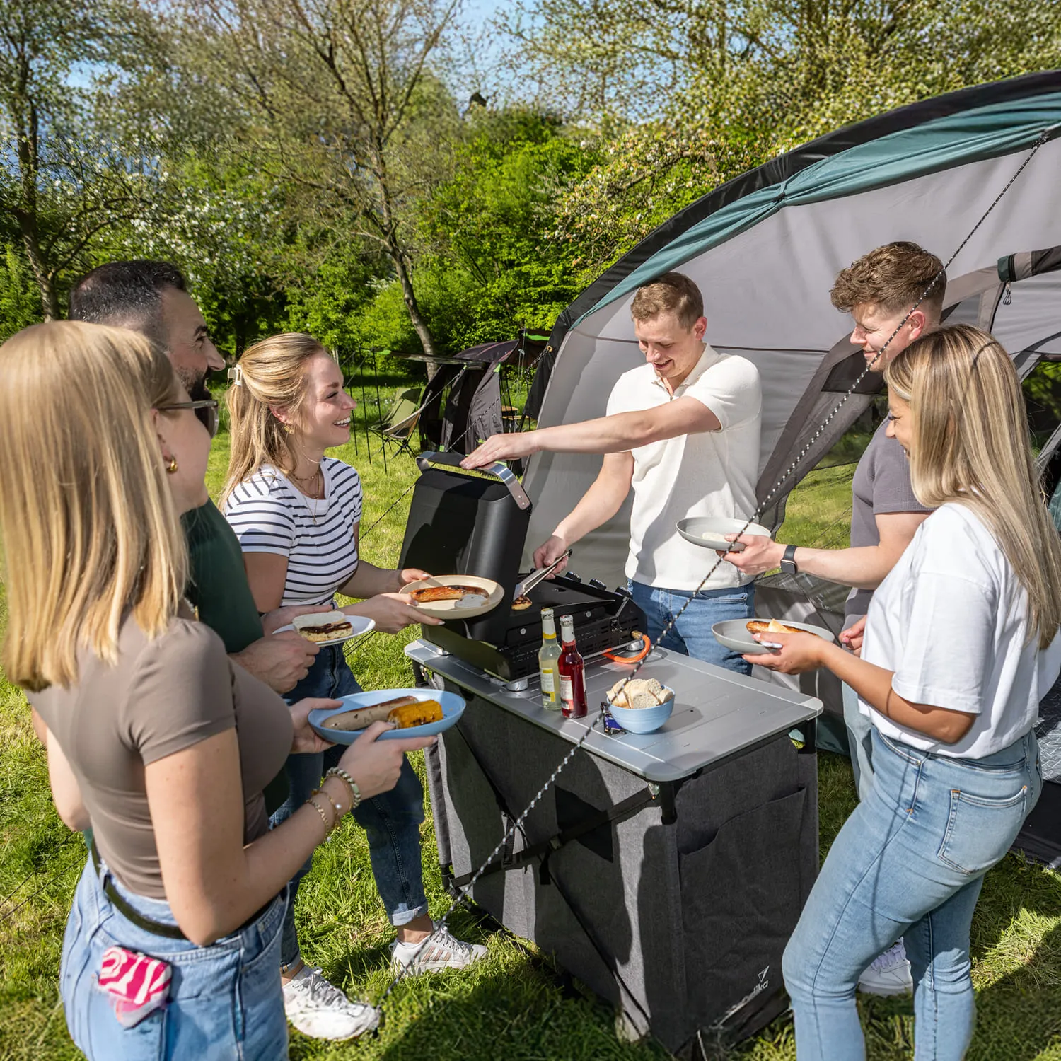 Grillstation im Freien mit sechs Personen, die Essen servieren und genießen. Zelt im Hintergrund, sonniger Tag.