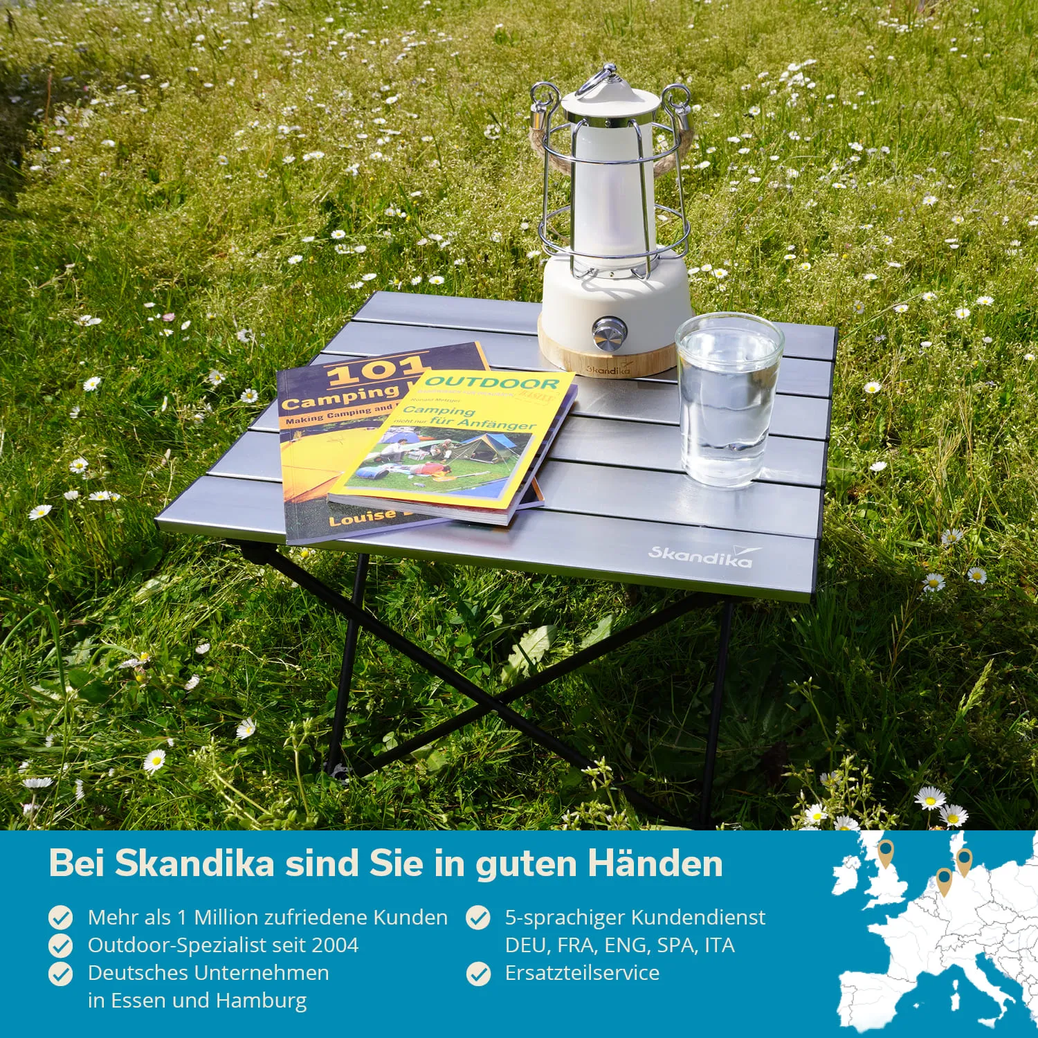 Ein klappbarer Tisch steht auf einer Wiese mit Blumen. Auf dem Tisch liegen zwei Campingbücher, eine weiße Laterne und ein Glas Wasser.