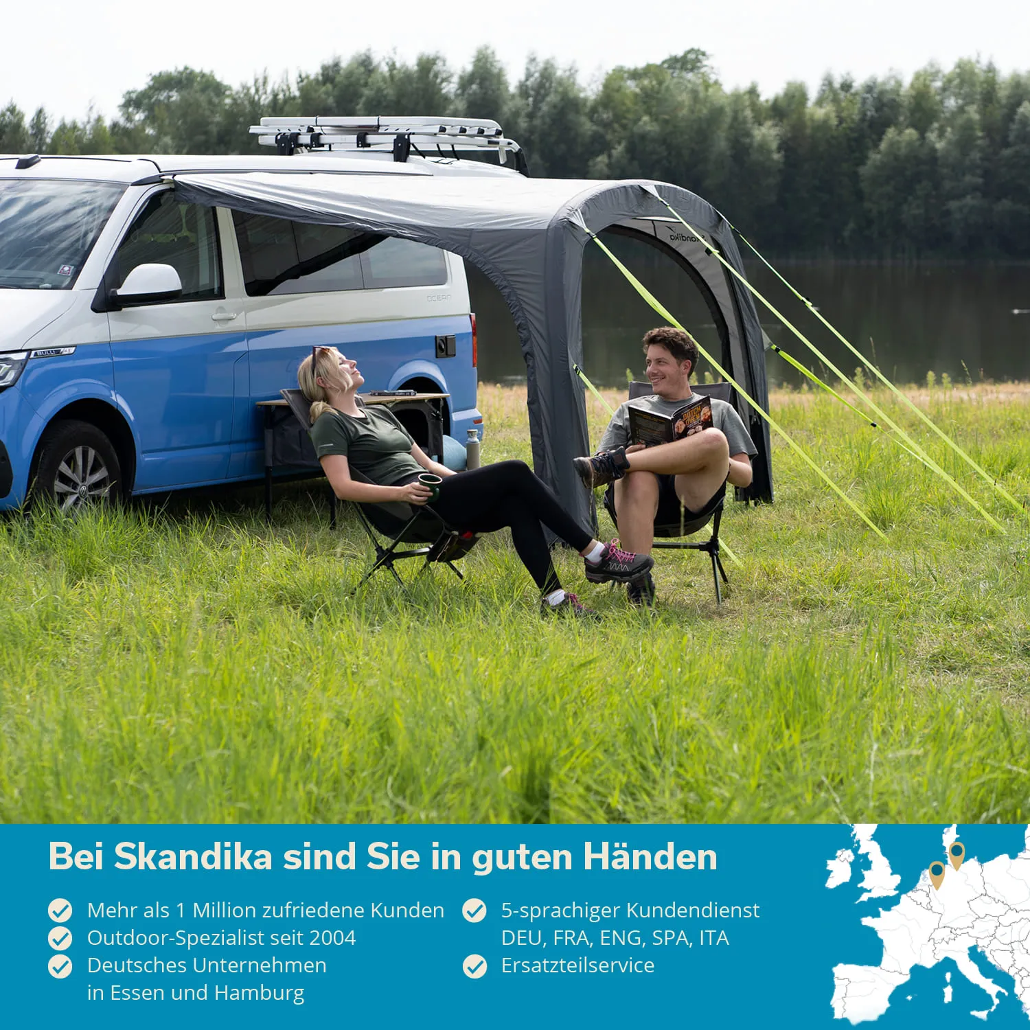 Zwei Personen sitzen auf Klappstühlen neben einem blauen Wohnmobil unter einem Gestell. Im Hintergrund gibt es Wasser und Bäume.