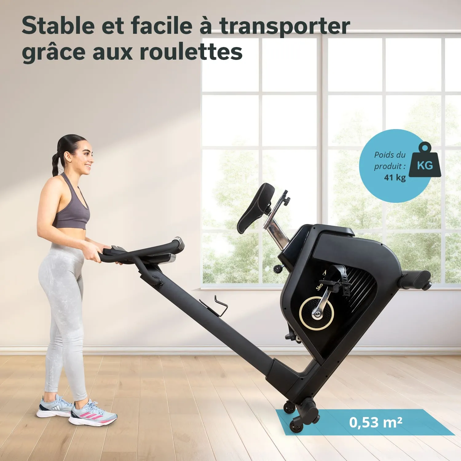 Ergometer in einem lichtdurchfluteten Raum. Eine Person hält das Gerät, das auf Rollen steht. Details wie Gewicht und Fläche sind sichtbar.