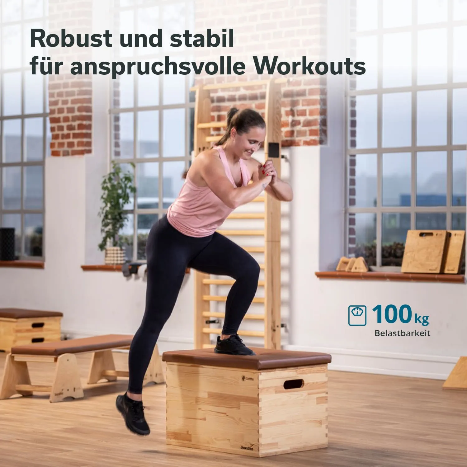 Eine Person steht auf einer Holzbank und führt eine Übung aus. Im Hintergrund sind Fenster und ein weiterer Trainingsbereich sichtbar.