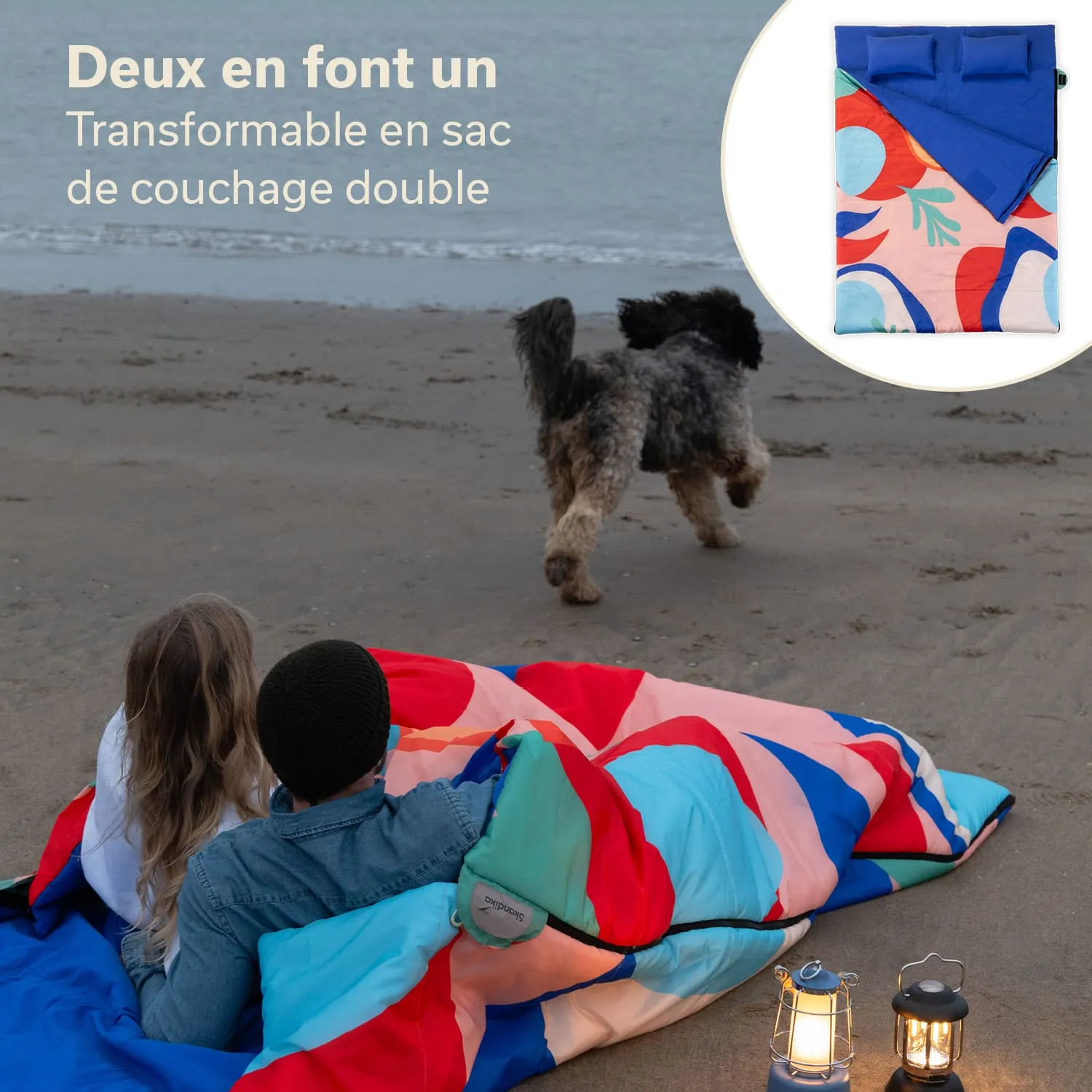 Eine Person sitzt mit einer weiteren Person auf einem Strand, beide in bunte Schlafsäcke gehüllt. Ein Hund läuft im Hintergrund. Es sind zwei Laternen zu sehen.