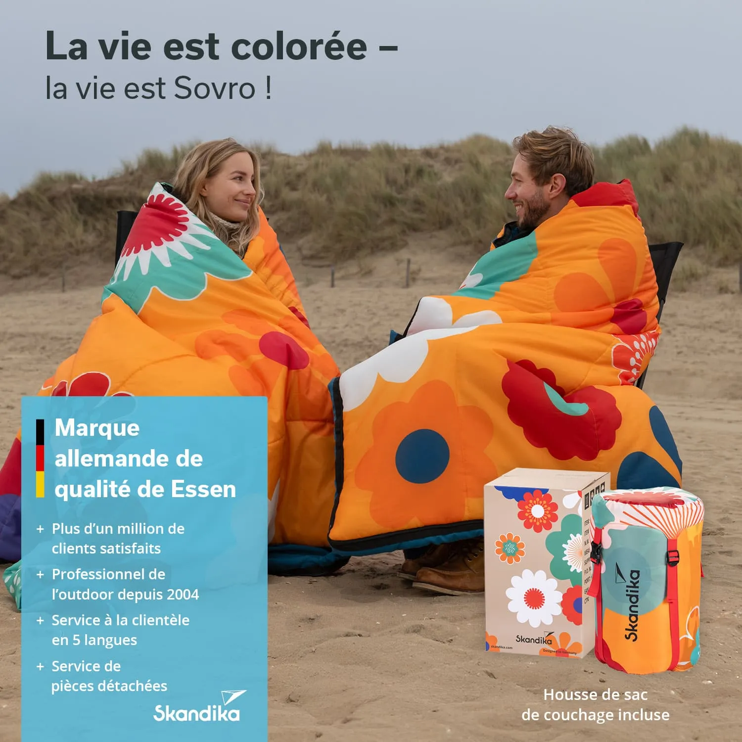 Zwei Personen sitzen im Sand am Strand, eingewickelt in bunte Decken mit Blumenmustern. Im Vordergrund stehen eine Verpackung und ein Kissen.