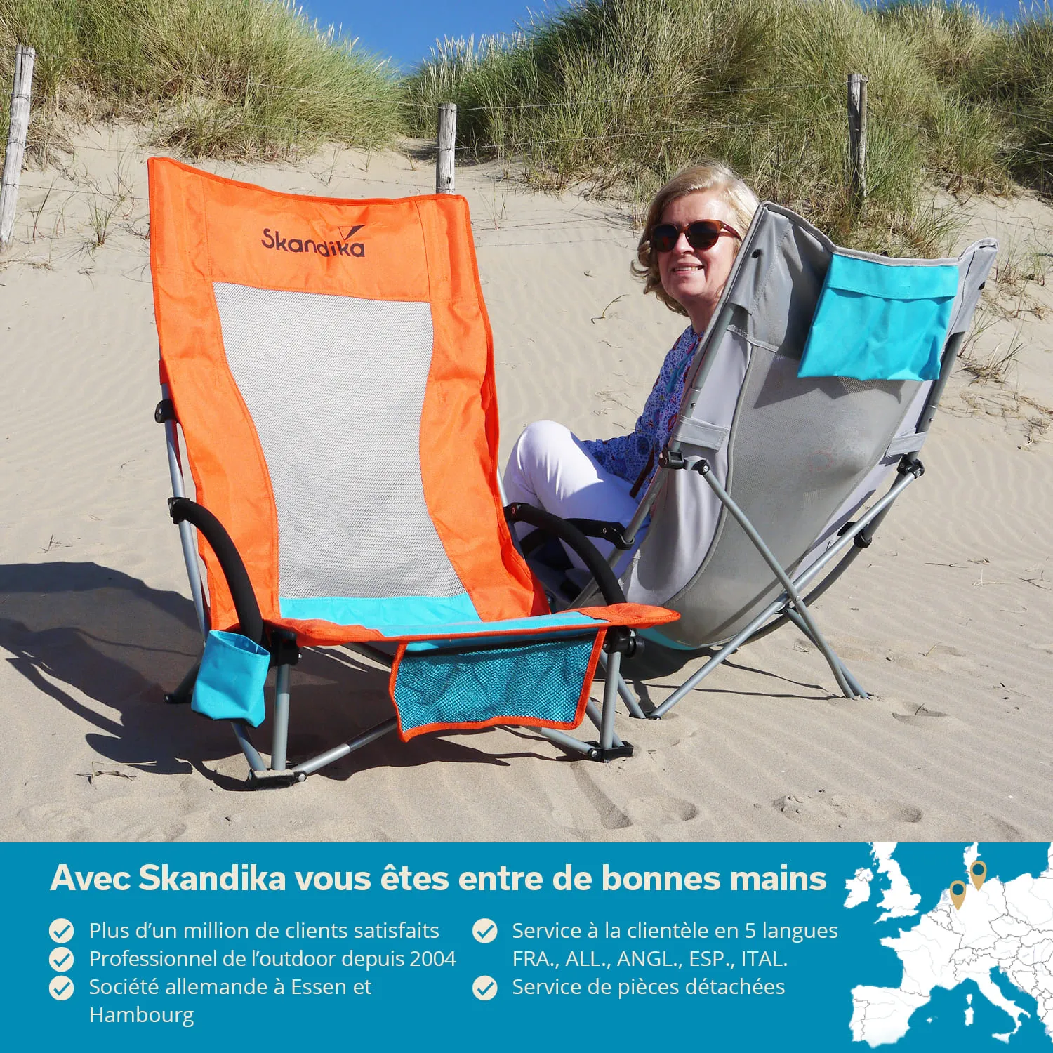 Zwei Klappstühle stehen im Sand, einer orange und einer grau. Eine Person sitzt in dem grauen Stuhl und lächelt. Grün bewachsene Dünen im Hintergrund.