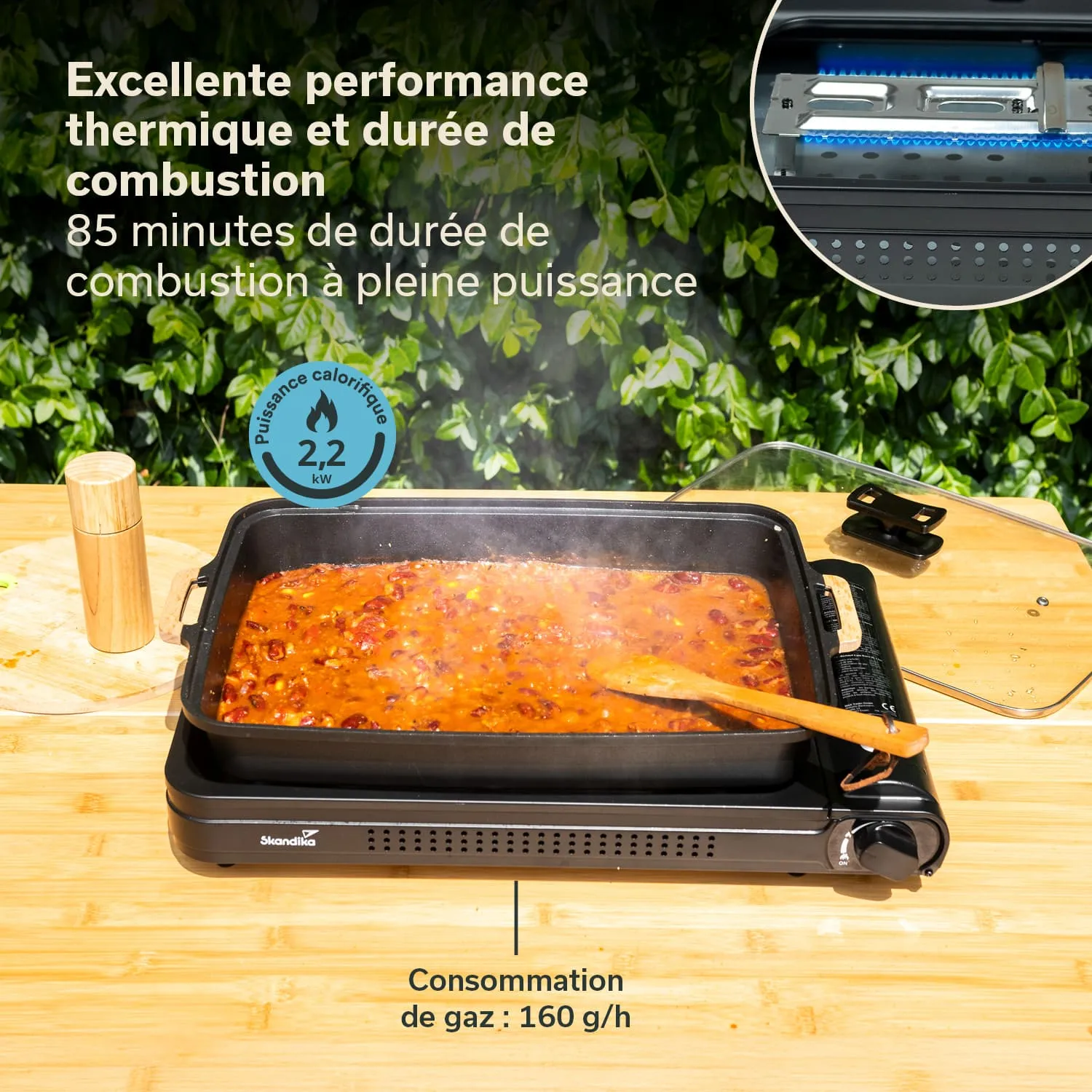 Ein elektrischer Grill mit einer Pfanne, in der eine dampfende, rotbraune Mischung kocht. Neben der Pfanne steht ein Holzsalzstreuer. Der Tisch ist aus Holz. Im Hintergrund sind grüne Pflanzenblätter sichtbar.