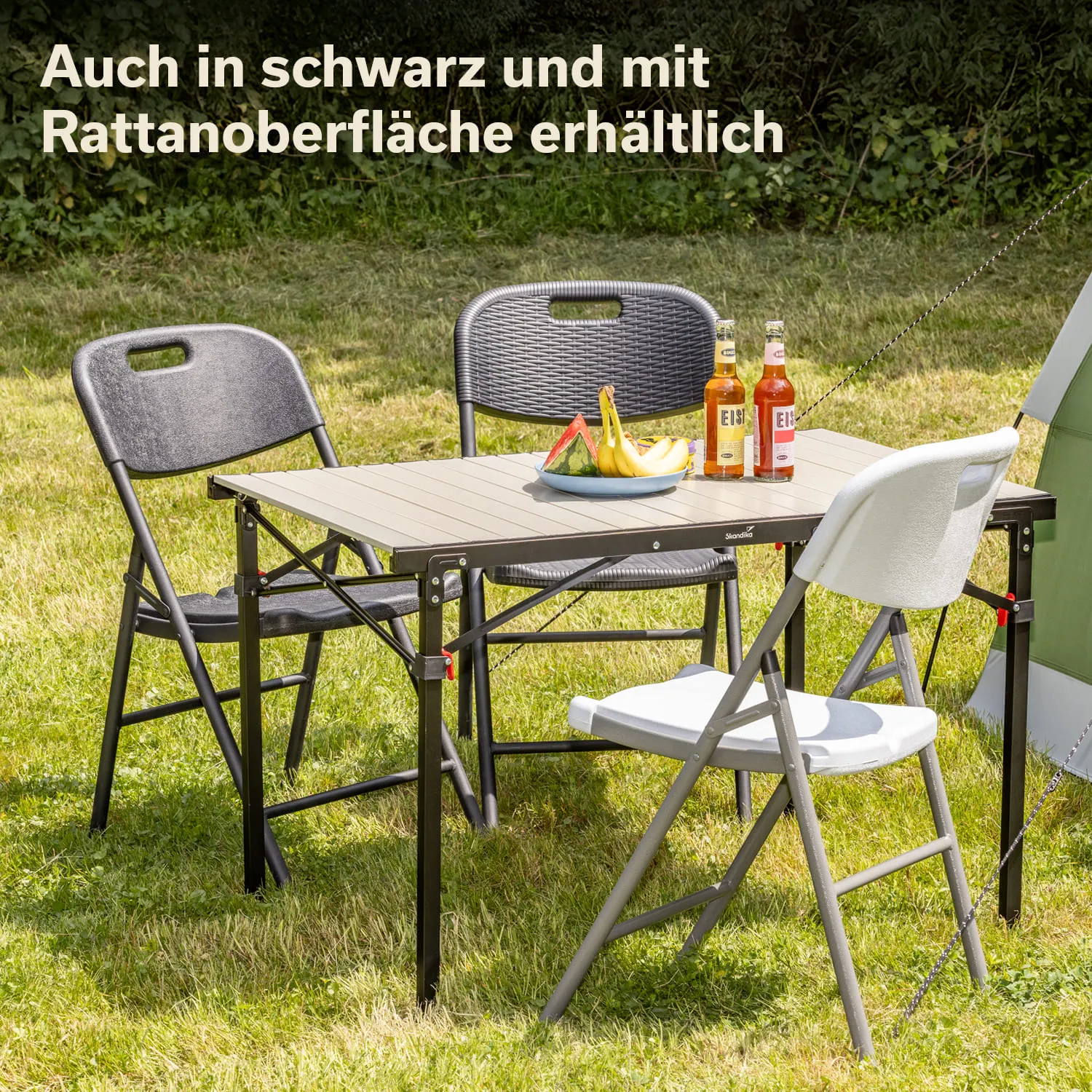 Tisch mit vier Klappstühlen in einem Grasbereich, Obst auf einem Teller und zwei Flaschen Getränke auf dem Tisch.
