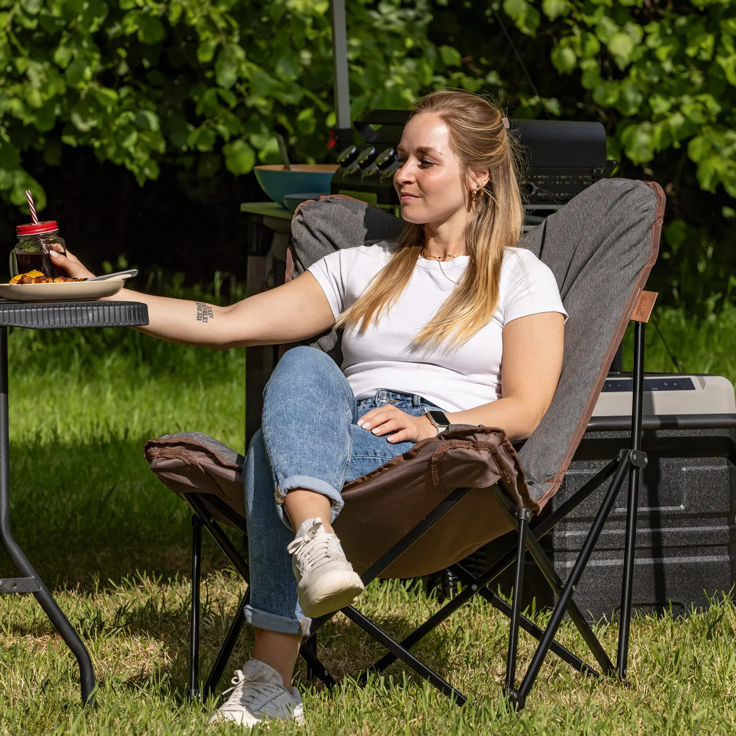 Person sitzt entspannt in einem Campingstuhl, hält einen Teller und ein Glas mit Strohhalm. Grünes Gras im Hintergrund.