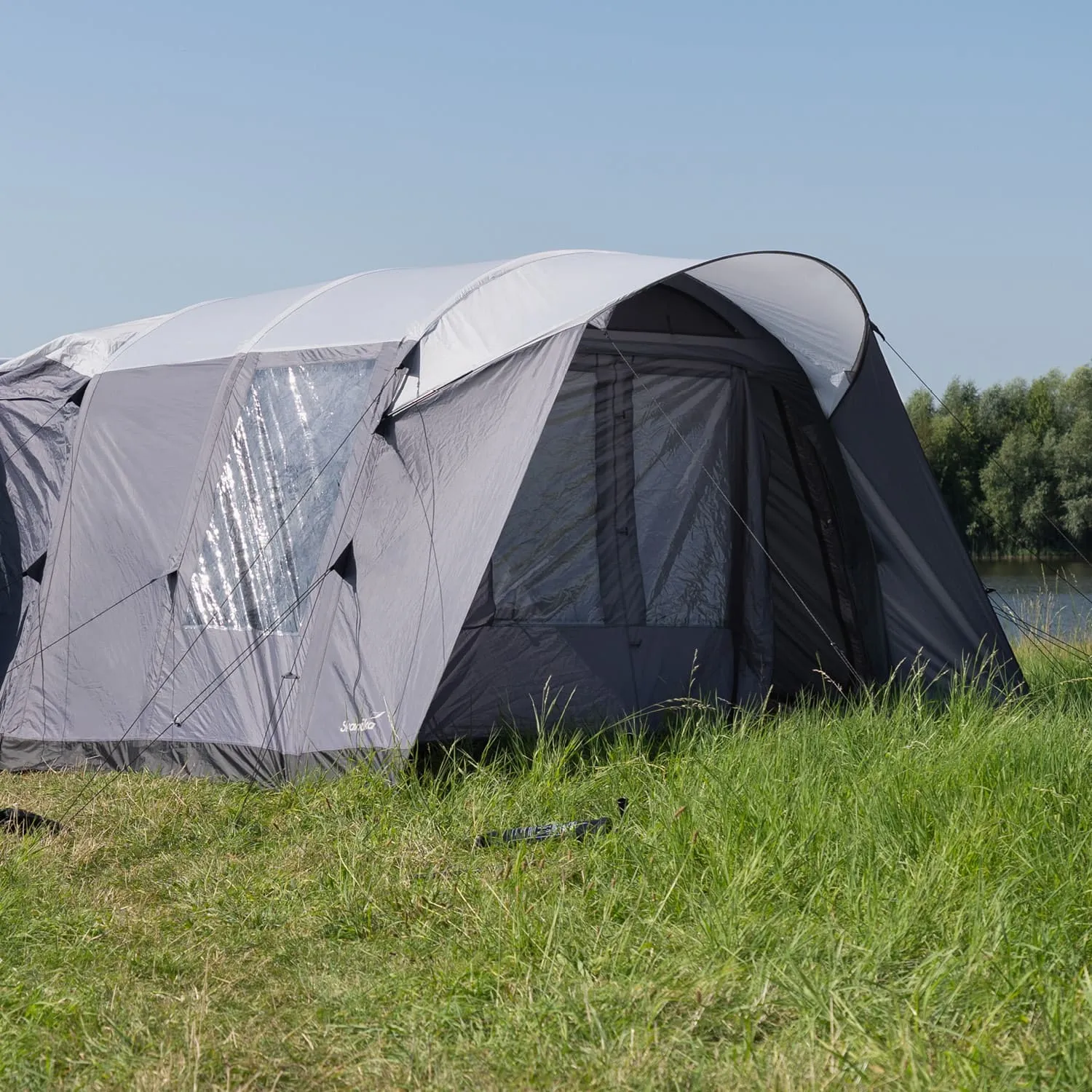 Graues Zelt mit transparenten Fenstern, auf einer grünen Wiese neben Gewässer, blauer Himmel im Hintergrund.