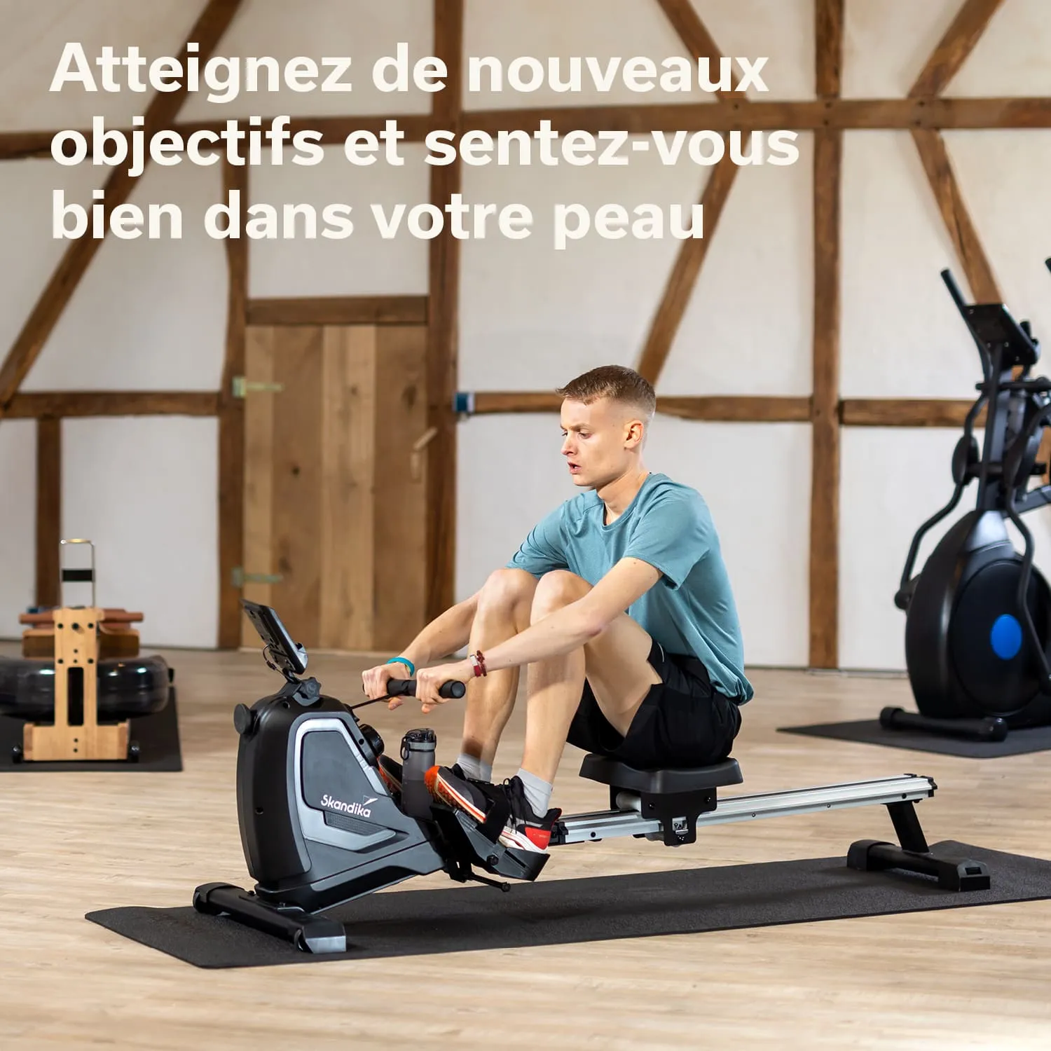 Person sitzt auf einem Rudergerät und trainiert in einem Raum mit Holzwänden. Im Hintergrund sind Fitnessgeräte sichtbar.