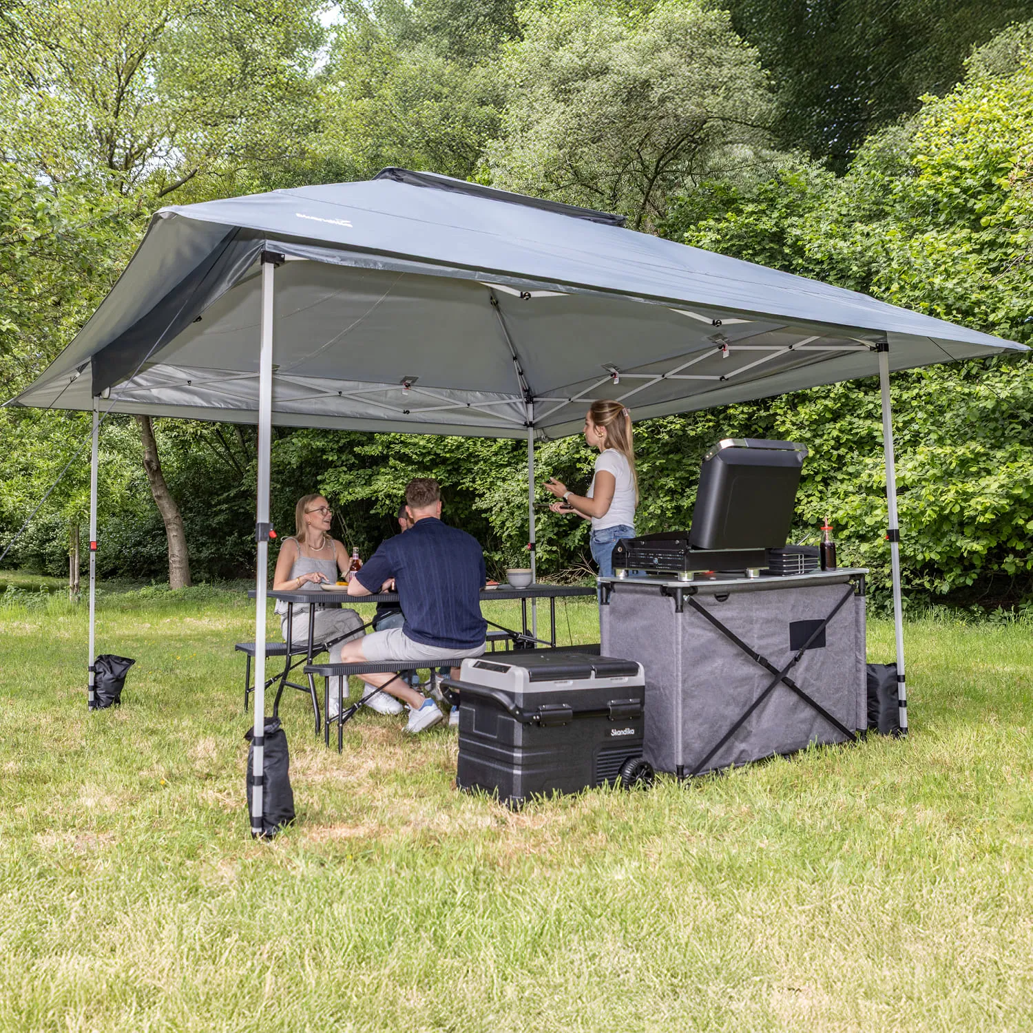 Zelt mit grauem Dach, darunter drei Personen an einem Tisch, eine weitere Person steht. Kühlbox und Grill sichtbar. Grünes Umfeld.