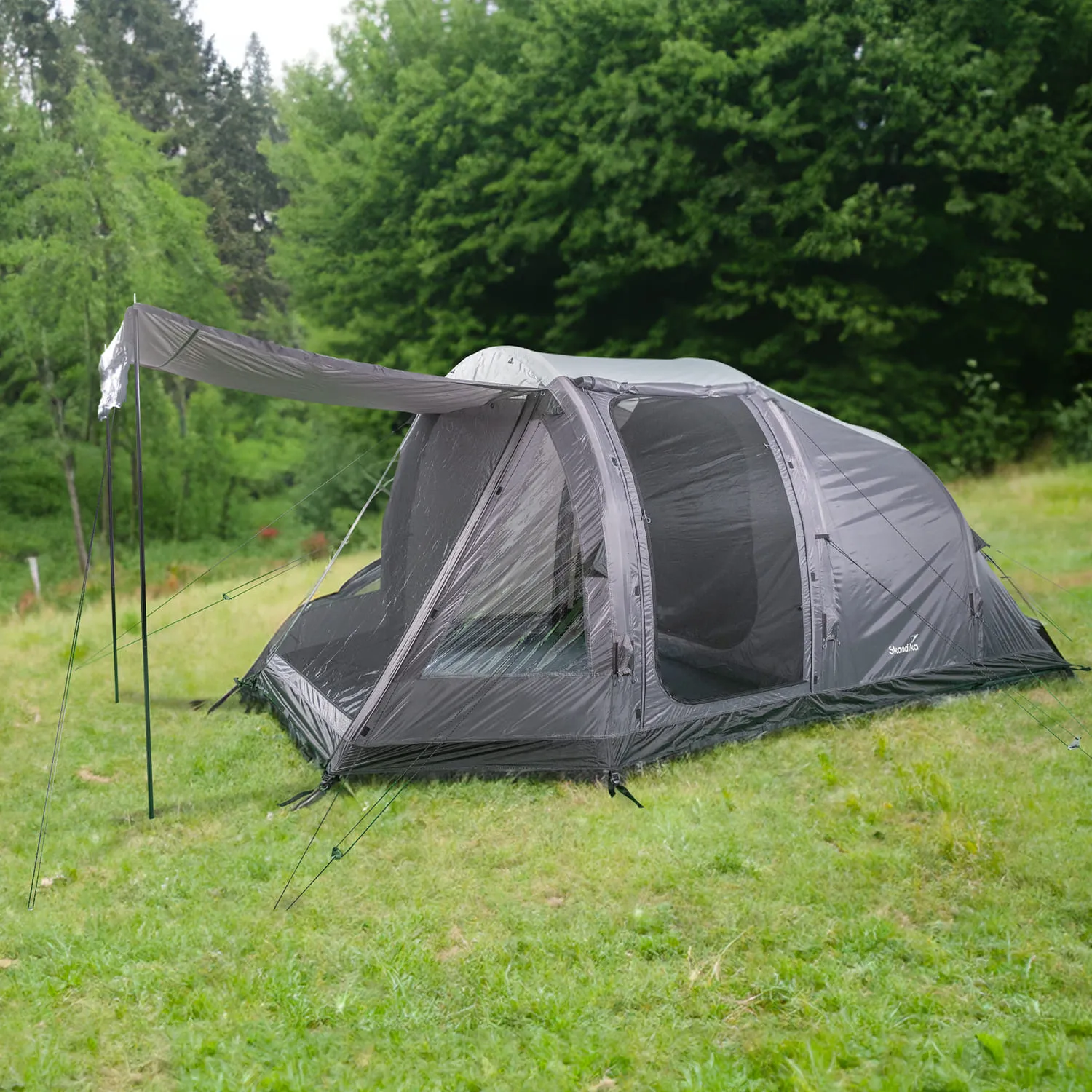 Grau-weißes Zelt auf einer grünen Wiese, umgeben von Bäumen. Ein großer Eingang mit Sichtschutz ist sichtbar.