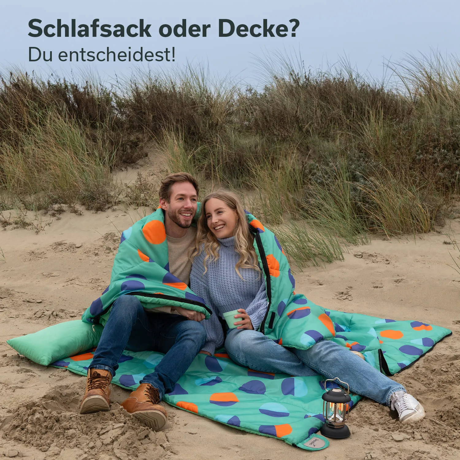 Paar sitzt auf einer Decke im Sand, eingekuschelt in einen bunten Schlafsack. Sie lächeln und halten eine Tasse. Hintergrund mit Schilf.