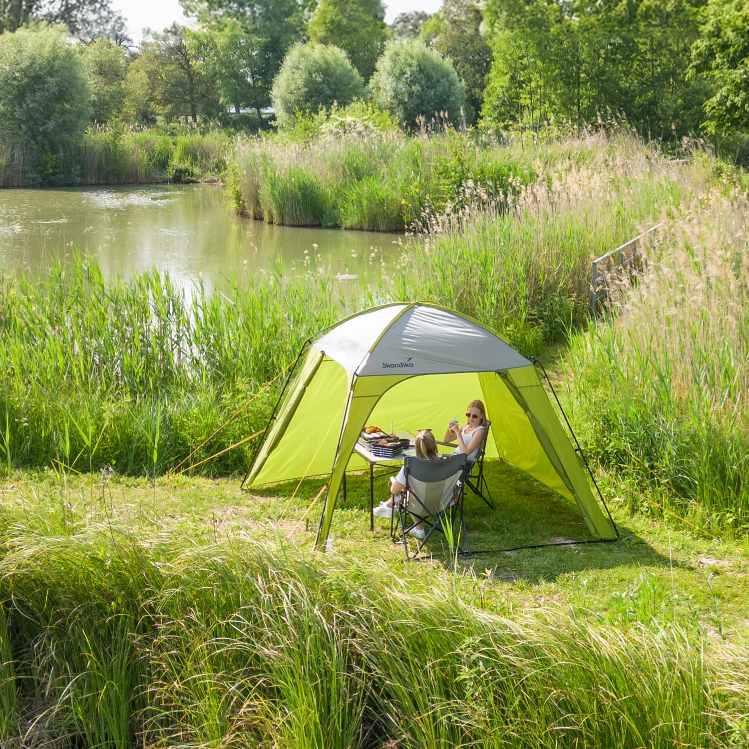 Zelt in der Natur am Ufer eines Gewässers, umgeben von hohen Gräsern. Zwei Personen sitzen entspannt am Tisch.
