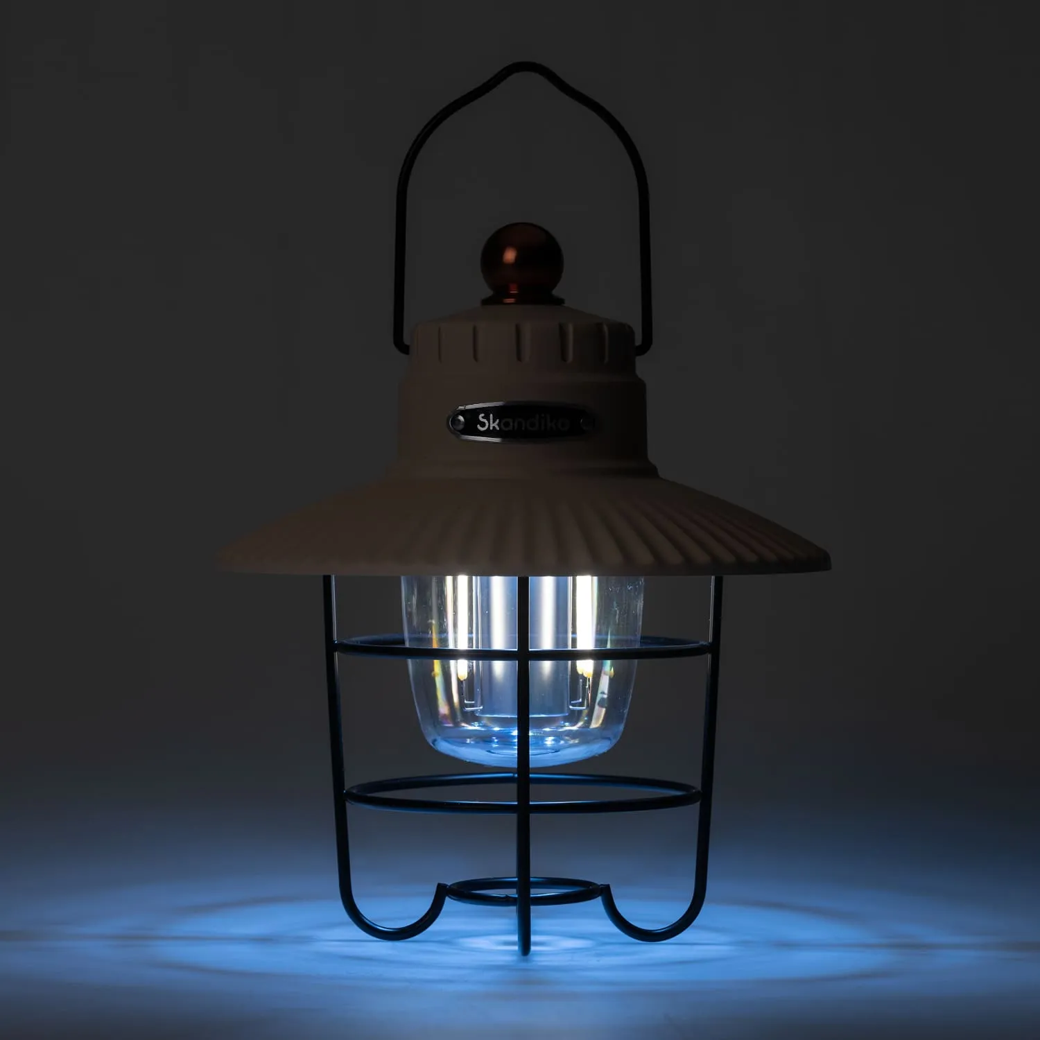 Eine hängende Öllampe mit einem runden, durchsichtigen Glaskörper, umgeben von einem schwarzen Metallgitter. Die Lampe hat einen beigen Schirm und eine kleine, runde Halterung oben. Der Hintergrund ist dunkel.