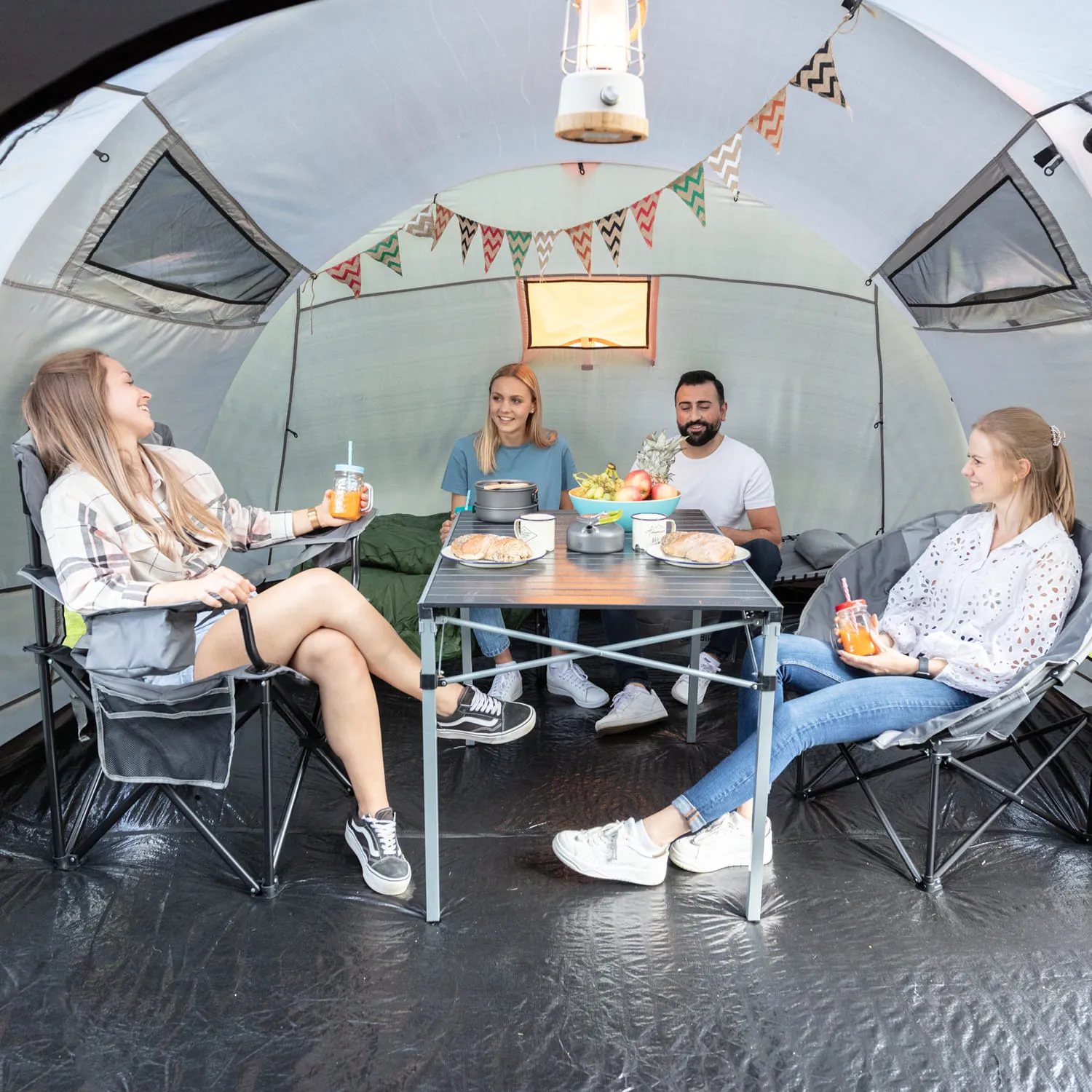 Vier Personen sitzen in einem Zelt um einen Tisch mit Snacks und Getränken. Bunte Wimpel sind dekorativ aufgehängt.