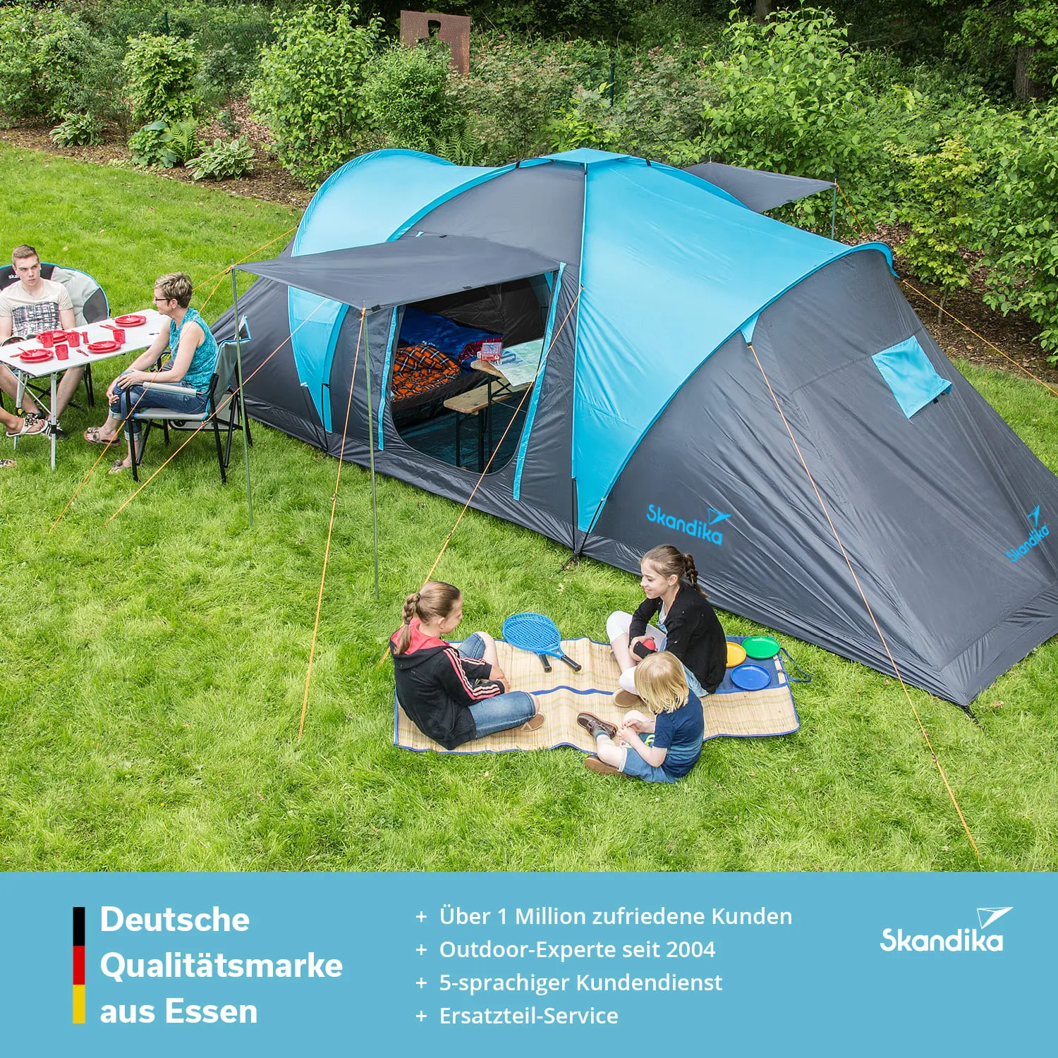 Zelt in der Natur mit mehreren Personen, die an einem Tisch sitzen und am Boden spielen. Grünes Gras und Bäume im Hintergrund.