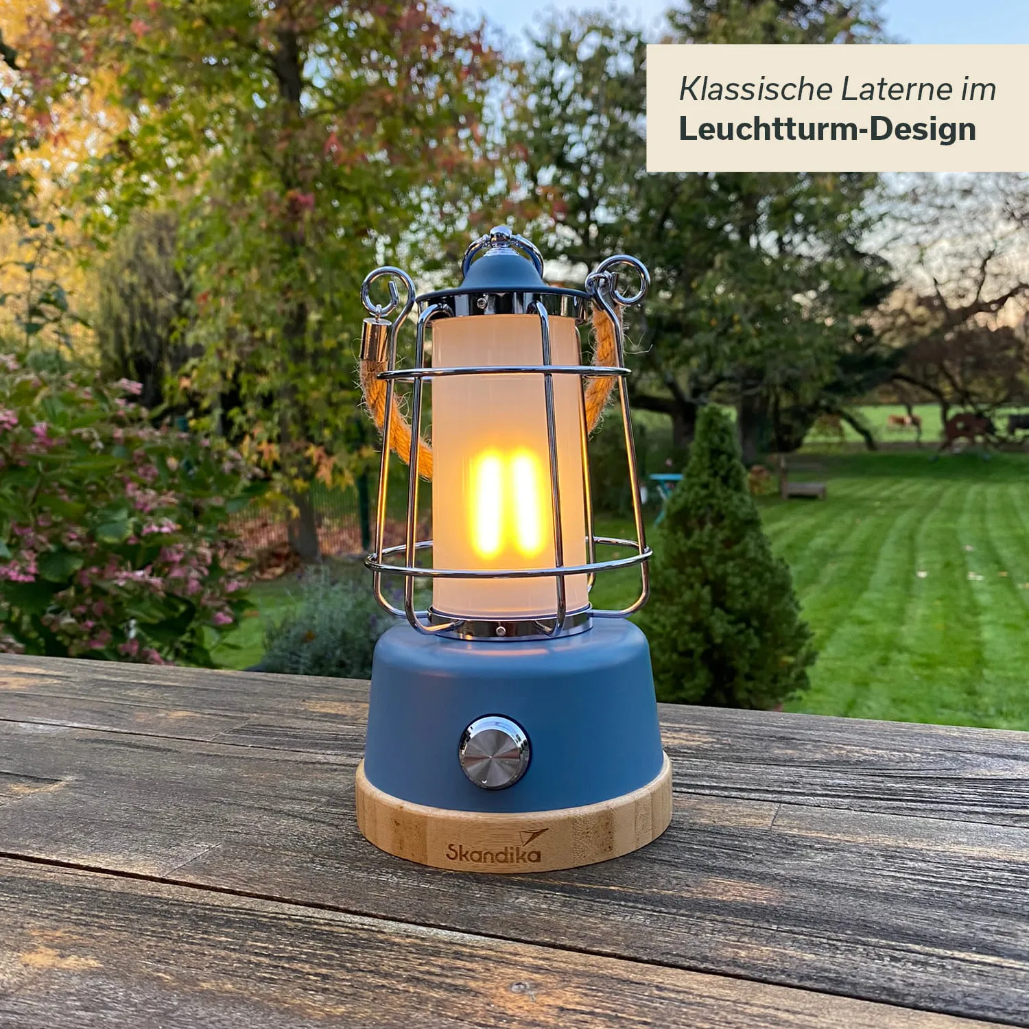 Eine klassische Laterne im Leuchtturm-Design steht auf einem Holztisch. Sie hat einen blauen Sockel mit einem Holzring und leuchtet. Im Hintergrund sind Bäume und ein gepflegter Rasen sichtbar.