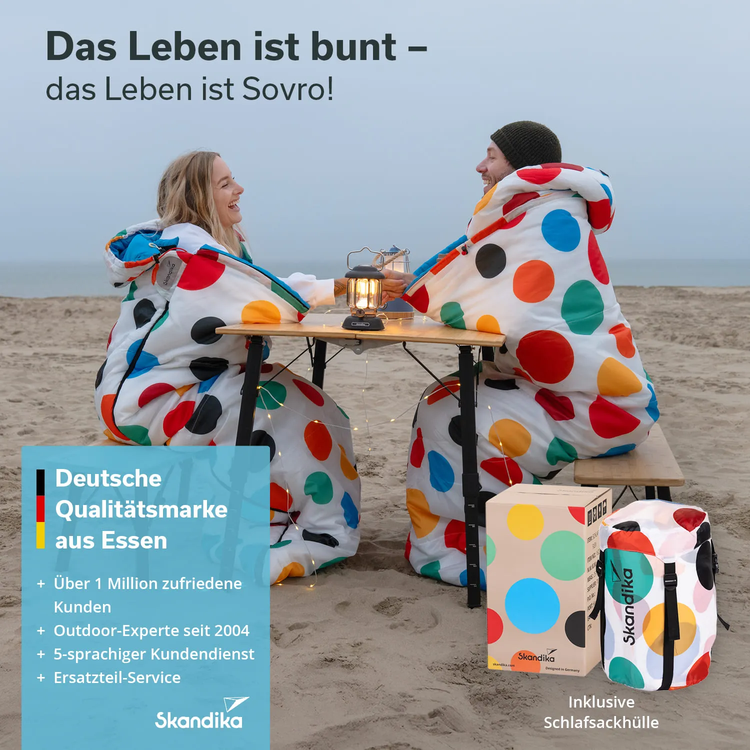 Zwei Personen in bunten Schlafsäcken sitzen an einem Tisch auf einem Strand. Bei Dämmerung, mit einer Laterne.
