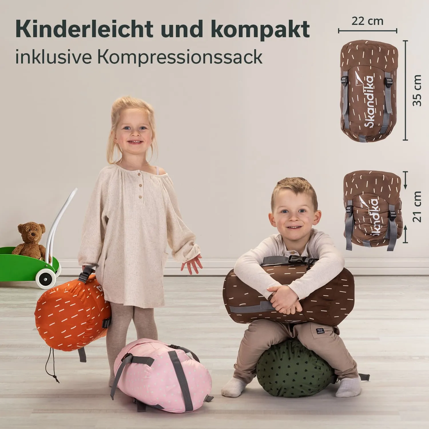 Zwei Kinder stehen in einem Raum mit hellen Wänden. Eines hält einen orangefarbenen, das andere einen braunen Rucksack. Auf dem Boden liegen ein pinker Rucksack und ein grüner Ball. Ein Spielzeugwagen mit einem Stoffbären ist im Hintergrund.