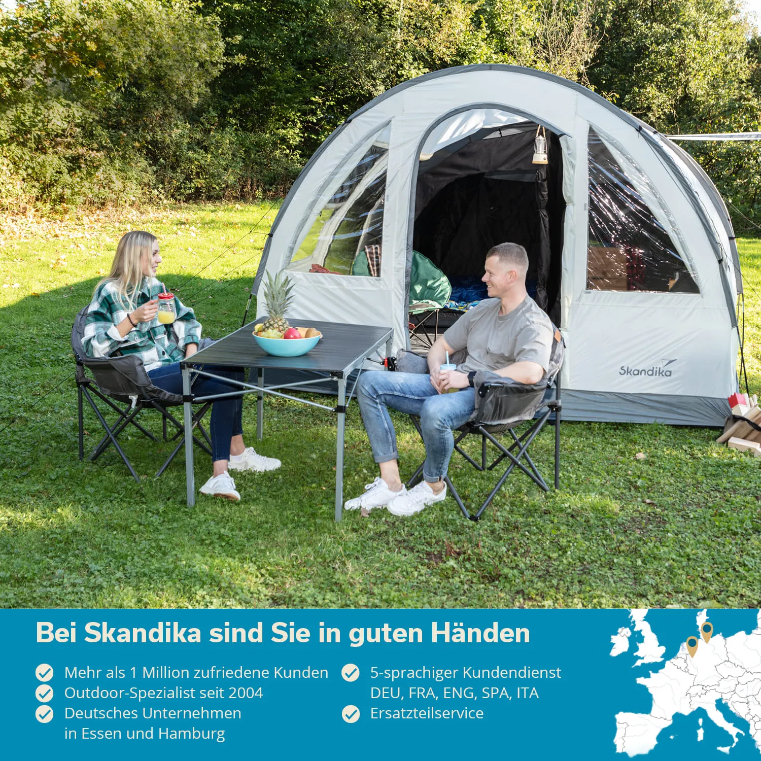 Zelt im Freien mit zwei Personen, die an einem Tisch mit Obst und Getränken sitzen. Grüner Rasen und Bäume im Hintergrund.