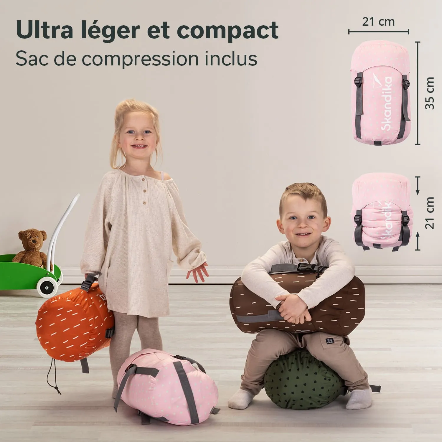Zwei Kinder stehen auf einem hellen Boden und halten farbige Taschen. Ein Kind sitzt auf einer runden, grünen Tasche. Im Hintergrund steht ein grünes Spielzeug mit einem Teddybären.