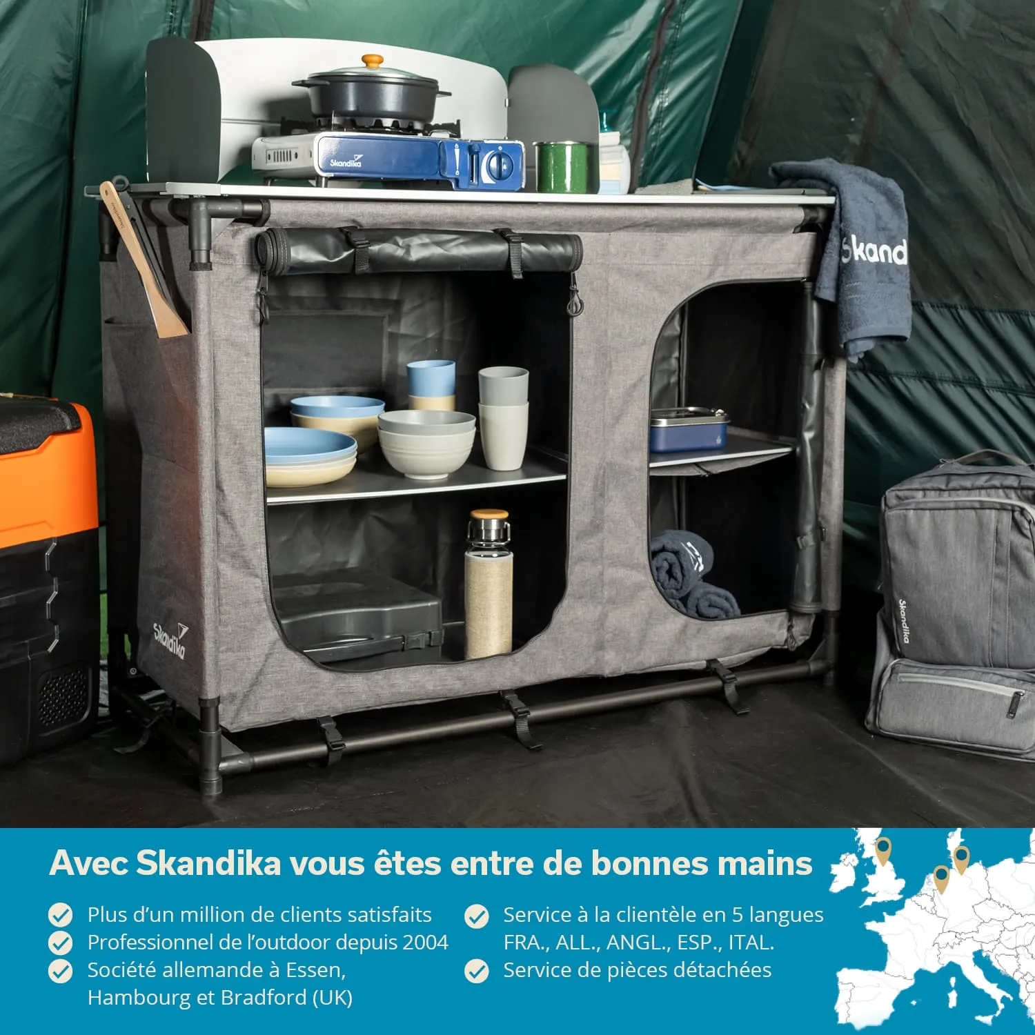 Eine graue Campingtisch-Einheit mit Kochplatte, oben Geschirr und Utensilien, darunter Rhyte bei Regalen und einem Rucksack.