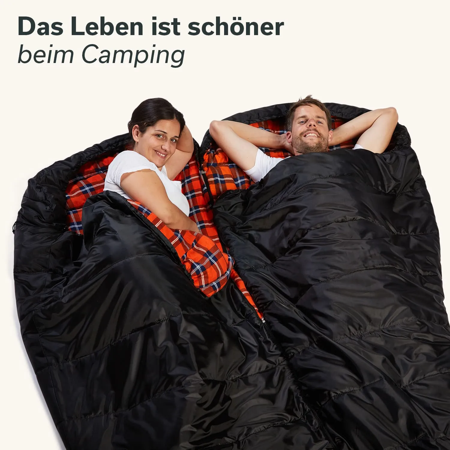 Zwei Personen sitzen in einem großen, schwarzen Schlafsack mit rotem Karomuster. Sie lächeln und lehnen sich entspannt zurück.