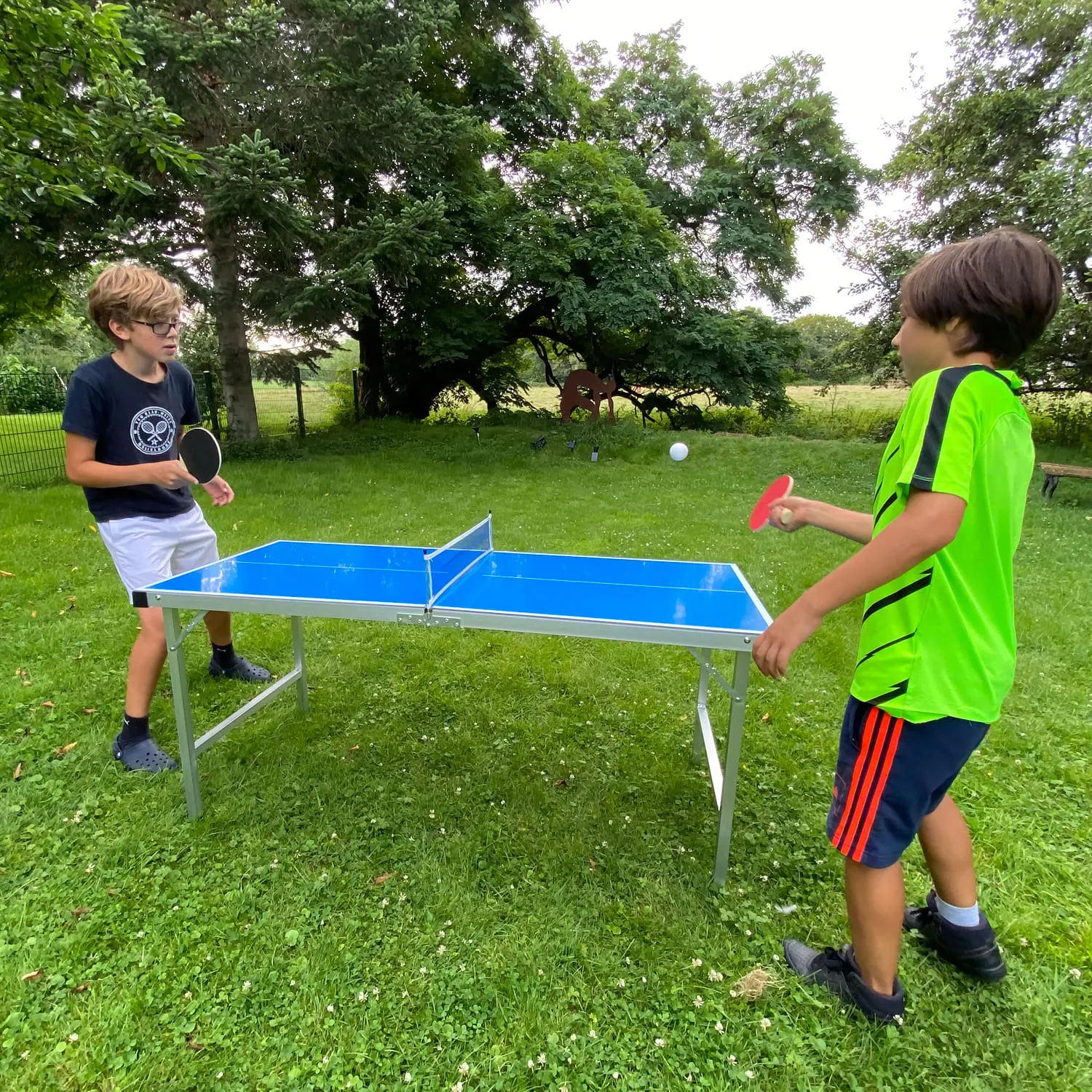 Zwei Personen stehen an einem blauen Tischtennistisch im Freien. Eine Person hält einen Tischtennisschläger, während die andere einen Ball schlägt. Im Hintergrund sind Bäume und ein weiteres Objekt sichtbar.