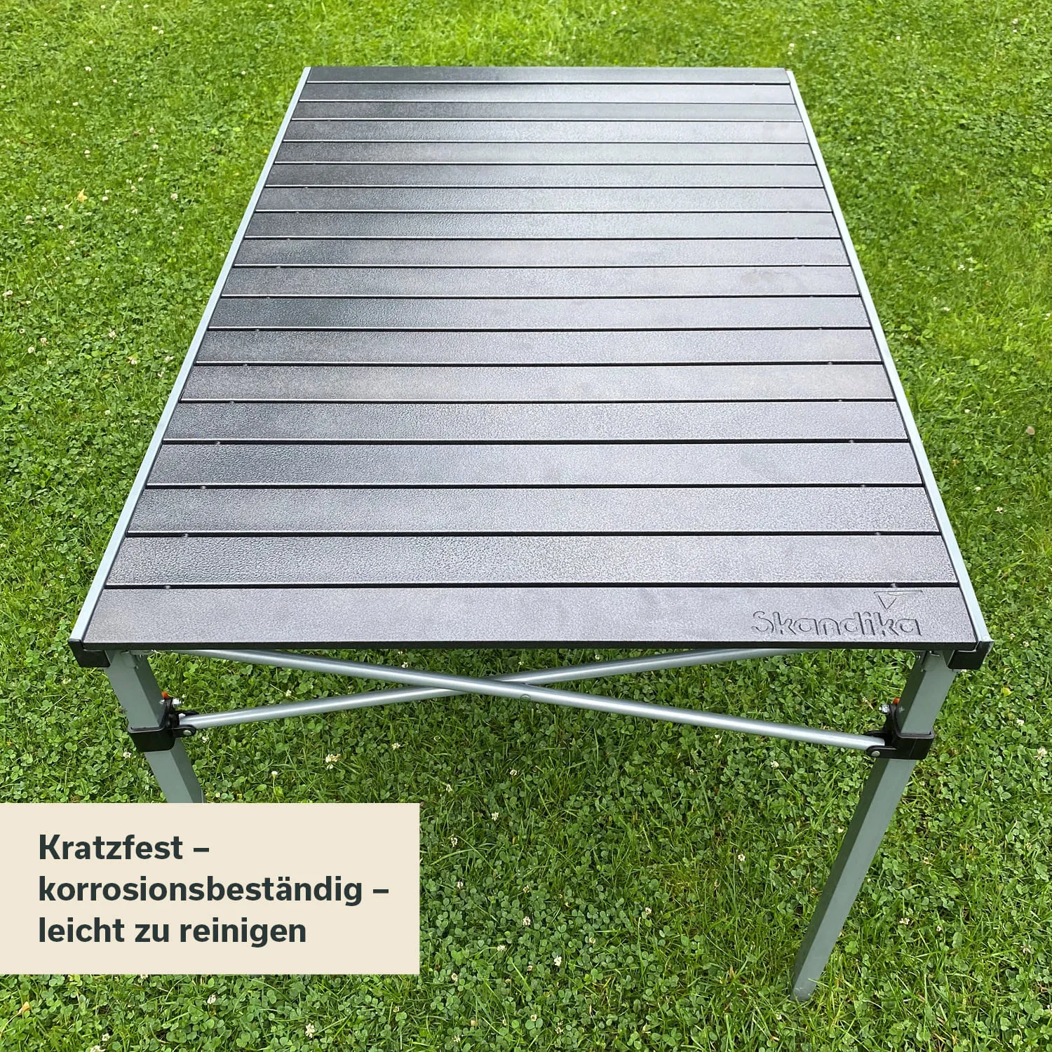 Ein grauer, klappbarer Tisch steht auf einer grünen Rasenfläche. Die Tischplatte hat eine gerillte Struktur.
