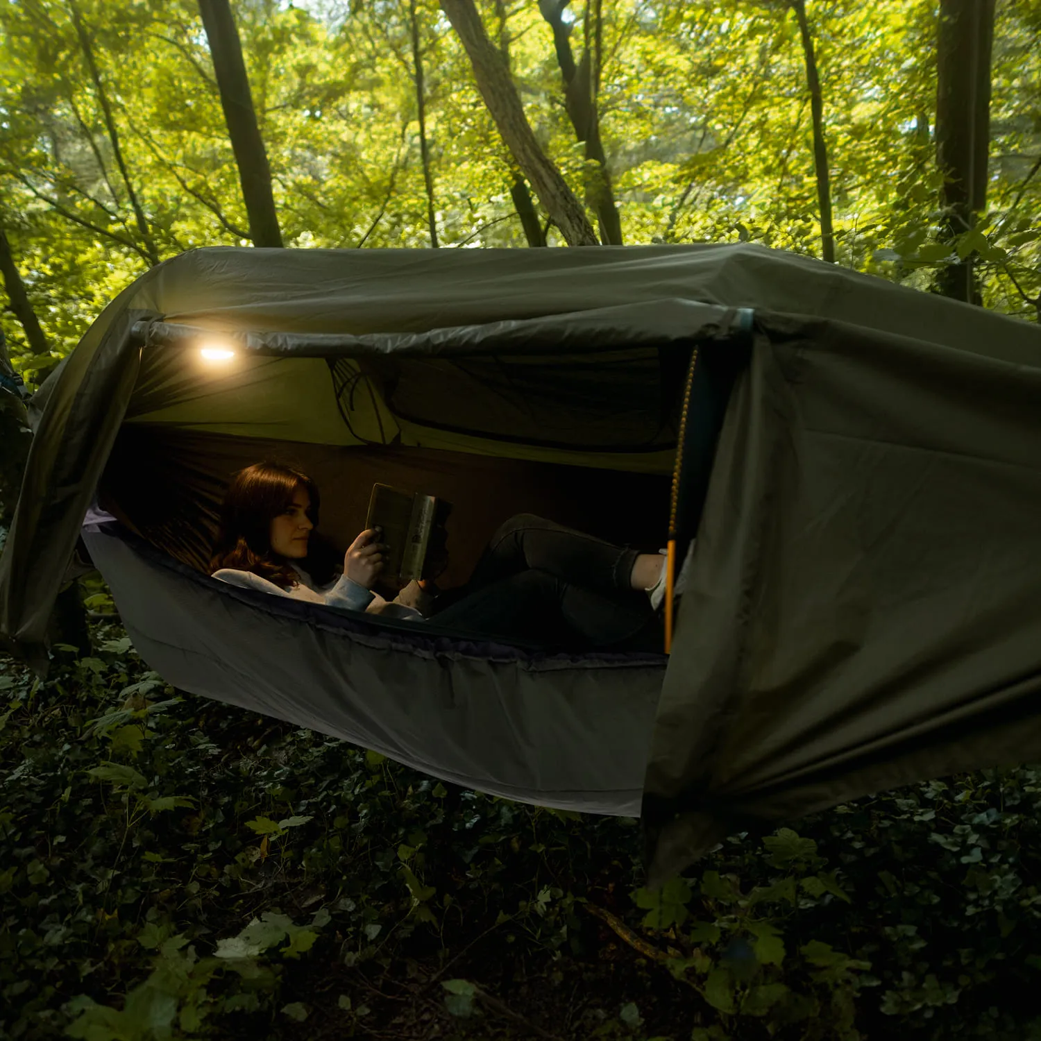 Person sitzt in einem hängenden Zelt im Wald und liest ein Buch. Umgeben von grünen Bäumen und Sträuchern.