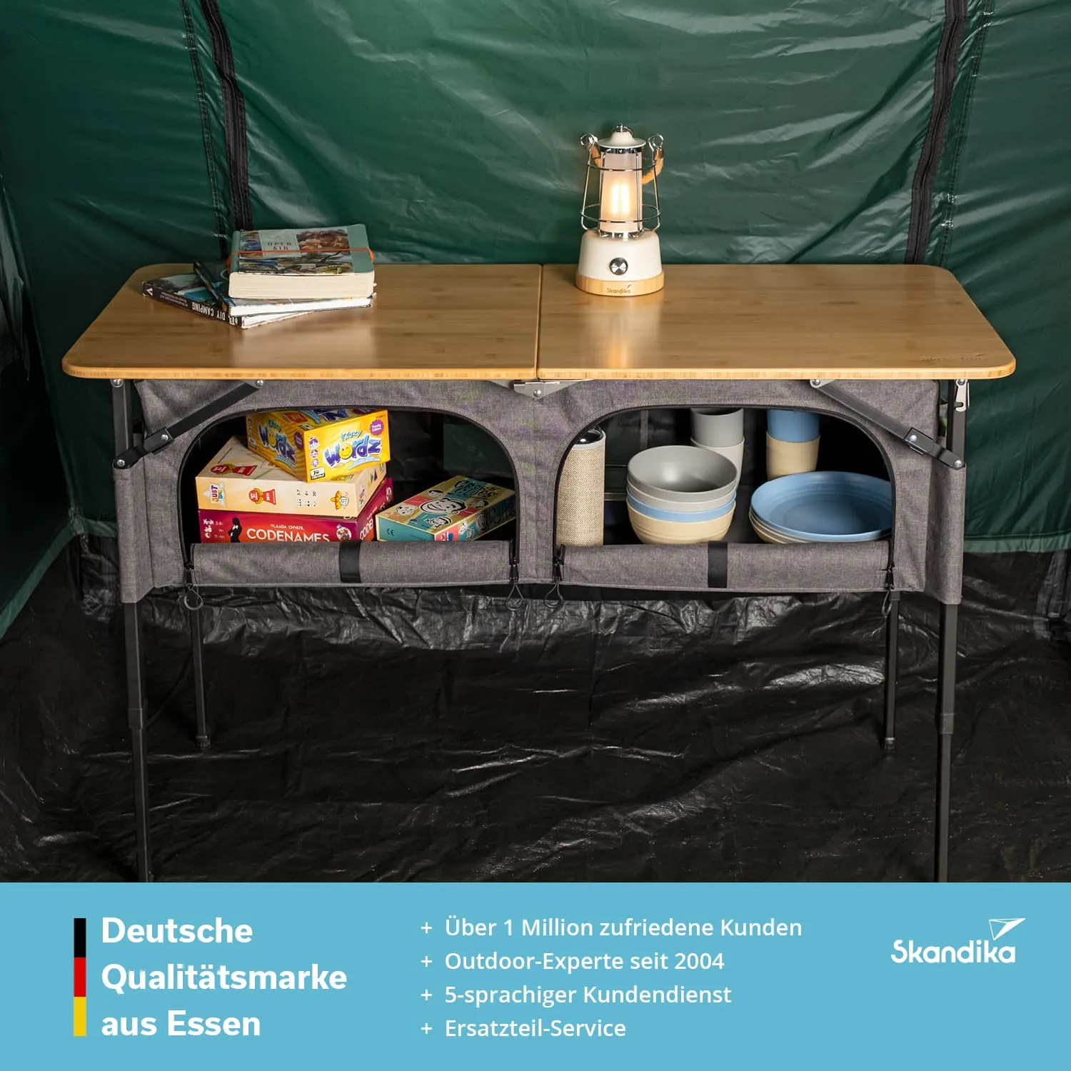 Ein klappbarer Campingtisch aus Holz mit einer schwarzen Unterlage. Auf dem Tisch befinden sich Bücher, ein Tischlicht und verschiedene Geschirrteile. Unter dem Tisch sind Spiele und Behälter sichtbar. Der Hintergrund ist grün.