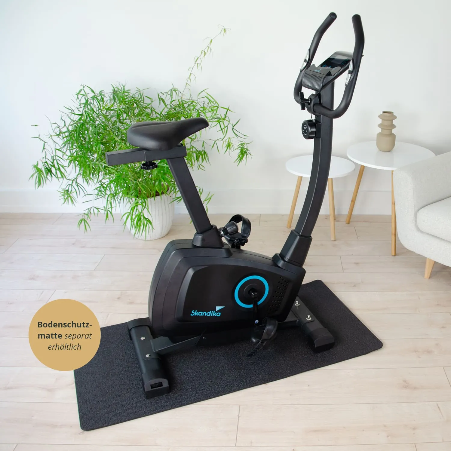 Schwarz-blauer Ergometer steht auf einer Bodenschutzmatte in einem modernen Raum mit Pflanze, kleinen Tischen und einem Sessel.