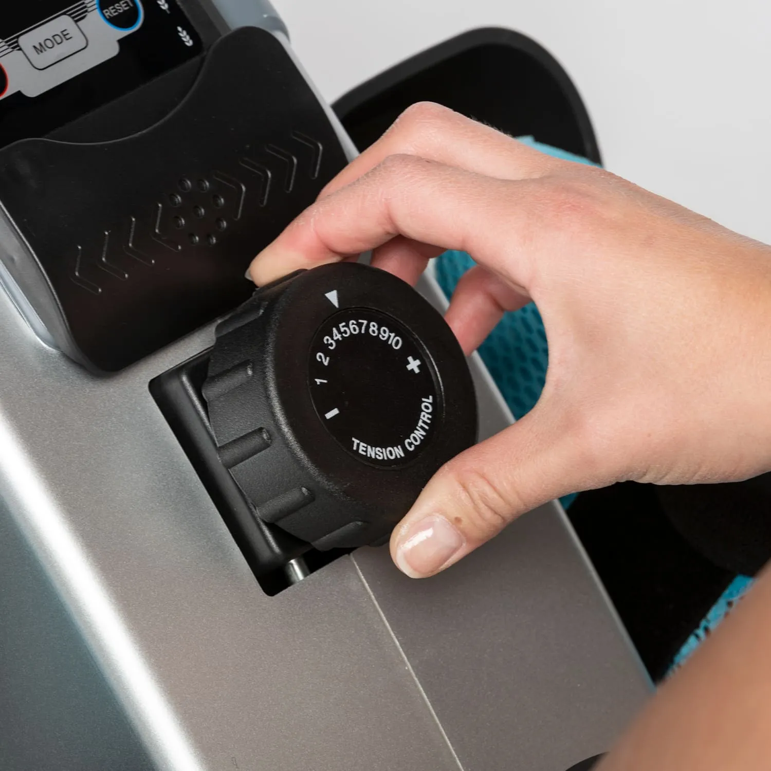 Hand dreht einen schwarzen Spannungsregler an einem Fitnessgerät. Das Gerät hat silberne und blaue Elemente.
