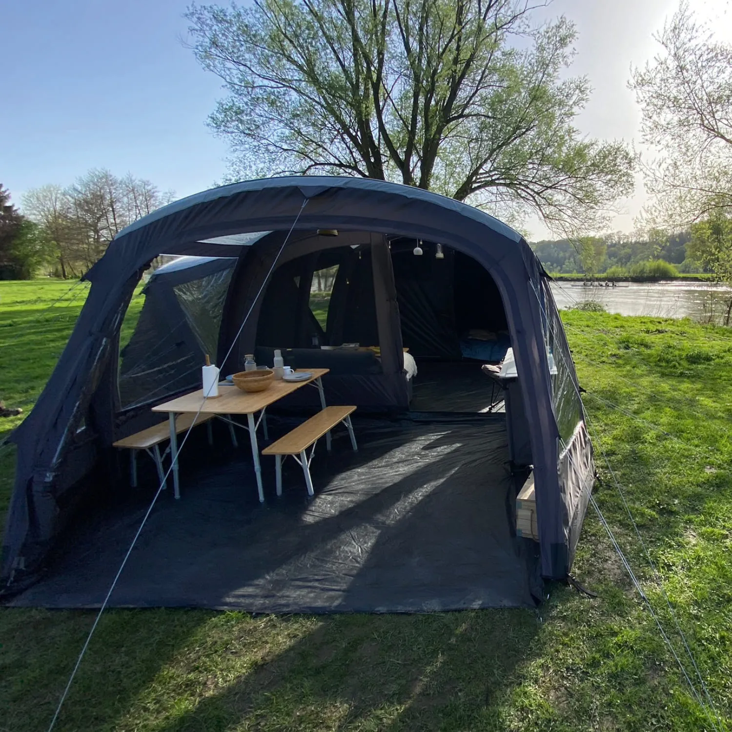 Zelt mit Tisch und Bänken auf einer Wiese, Blick auf einen Fluss und Bäume im Hintergrund.