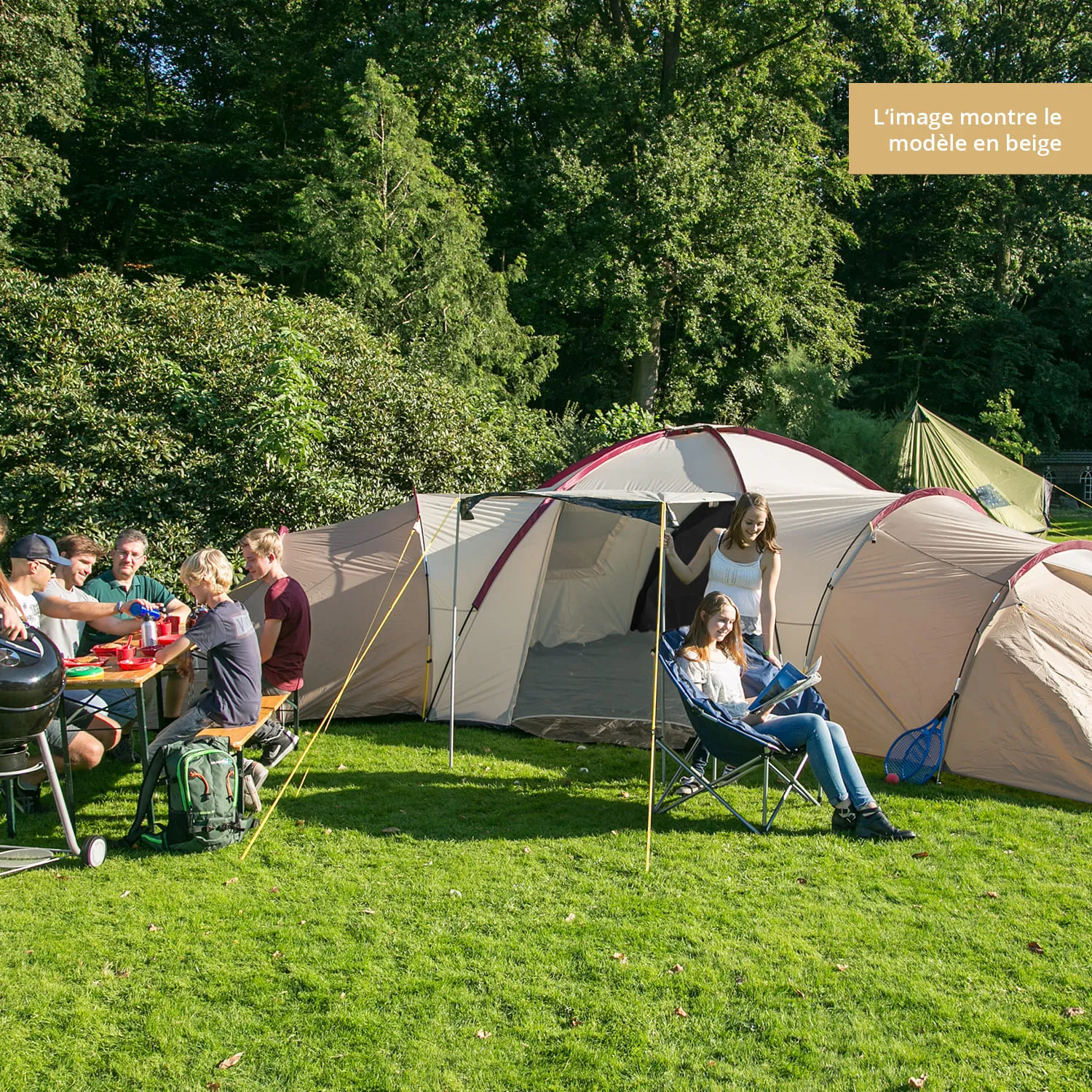 Eine große, helle Campingausrüstung auf Rasen, umgeben von Bäumen. Menschen sitzen an einem Tisch und essen, während andere entspannt auf Stühlen sitzen.