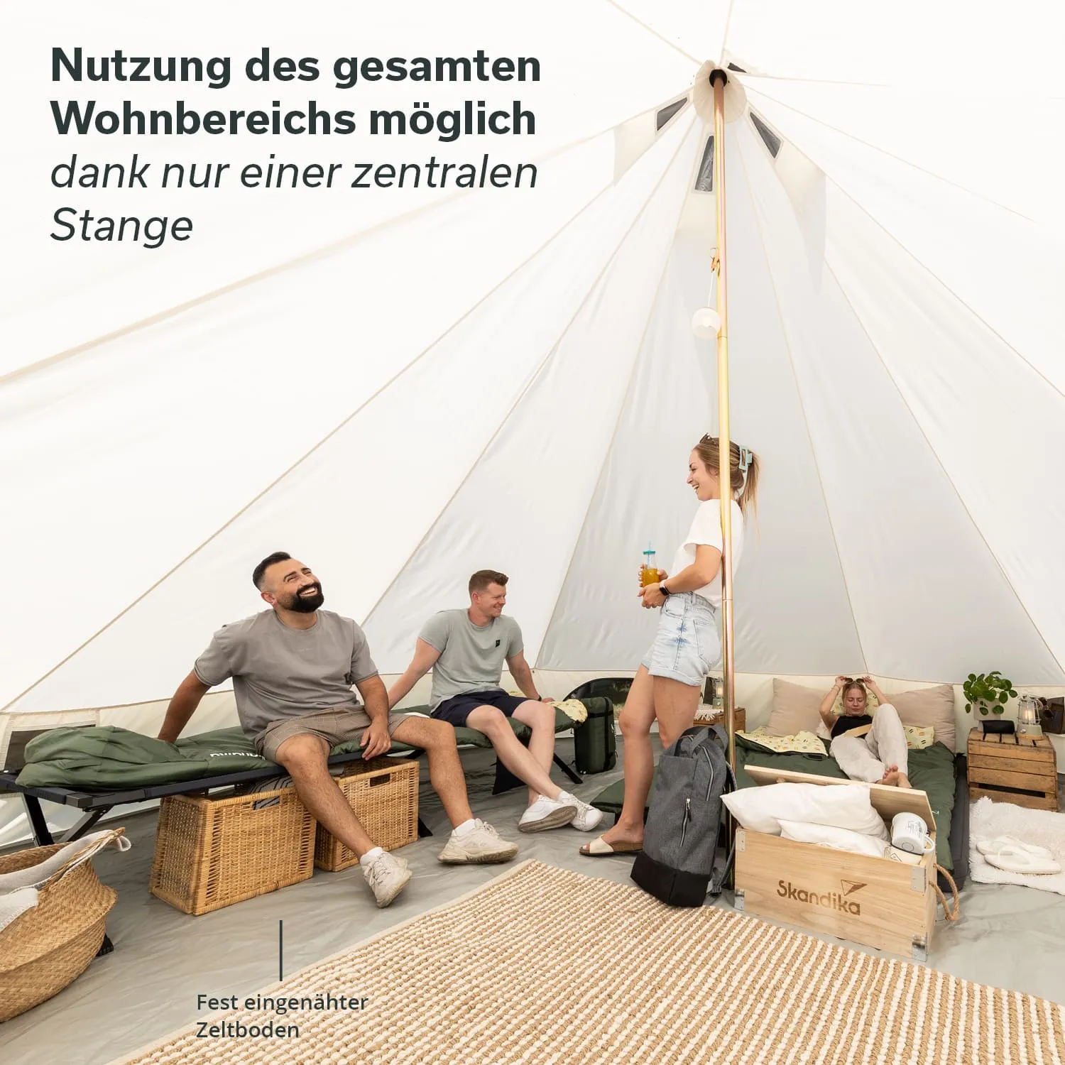 Innere eines großen Zeltes mit zentraler Stange. Vier Personen sitzen oder stehen, ein Handtuch und eine Pflanze sind sichtbar. Kenntnisse der Einrichtung.