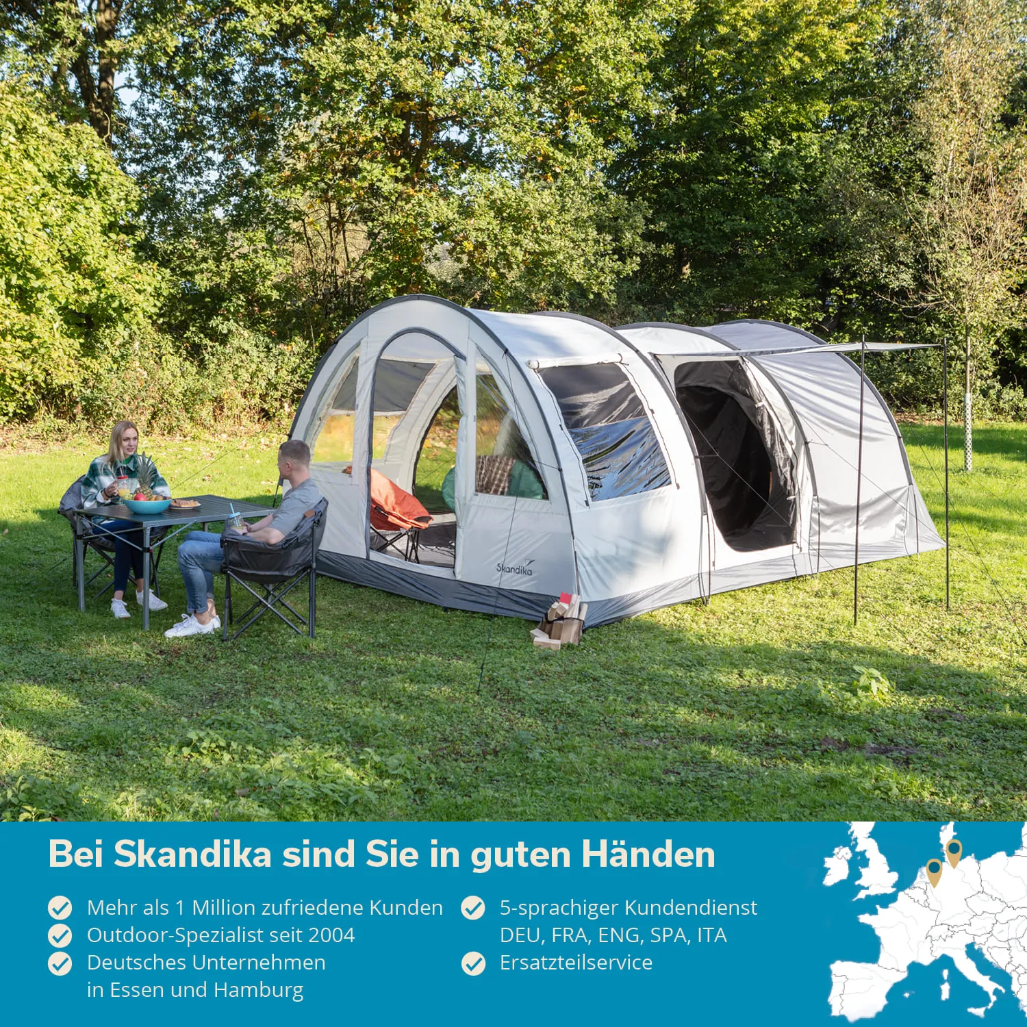 Zelt in einer grünen Wiese, zwei Personen sitzen am Tisch. Baumreiche Umgebung, ein Wäschegestell ist sichtbar.