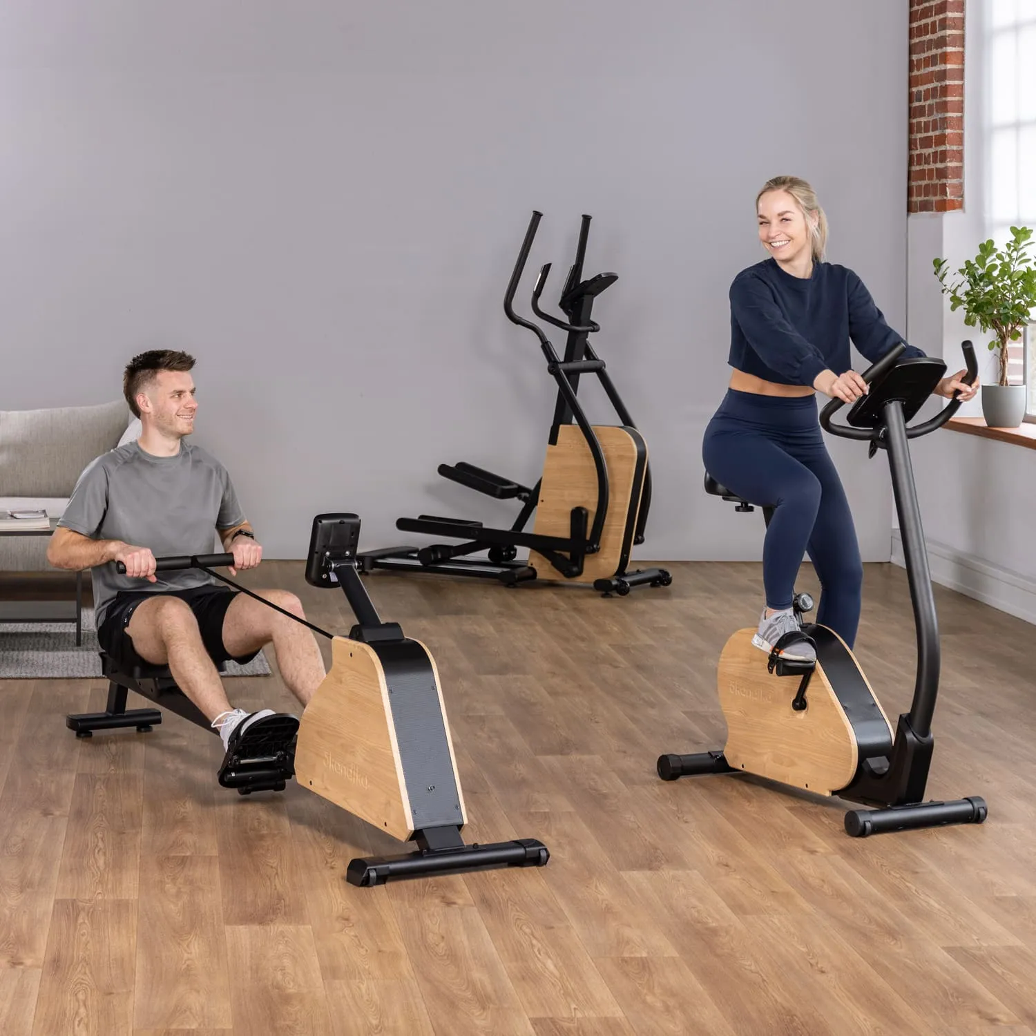 Zwei Personen trainieren in einem modernen Fitnessraum. Eine Person sitzt an einem Rudergerät, die andere fährt auf einem Heimtrainer. Beide Geräte haben Holz- und Metallverarbeitung. Im Hintergrund sind eine Couch und weitere Fitnessgeräte sichtbar.