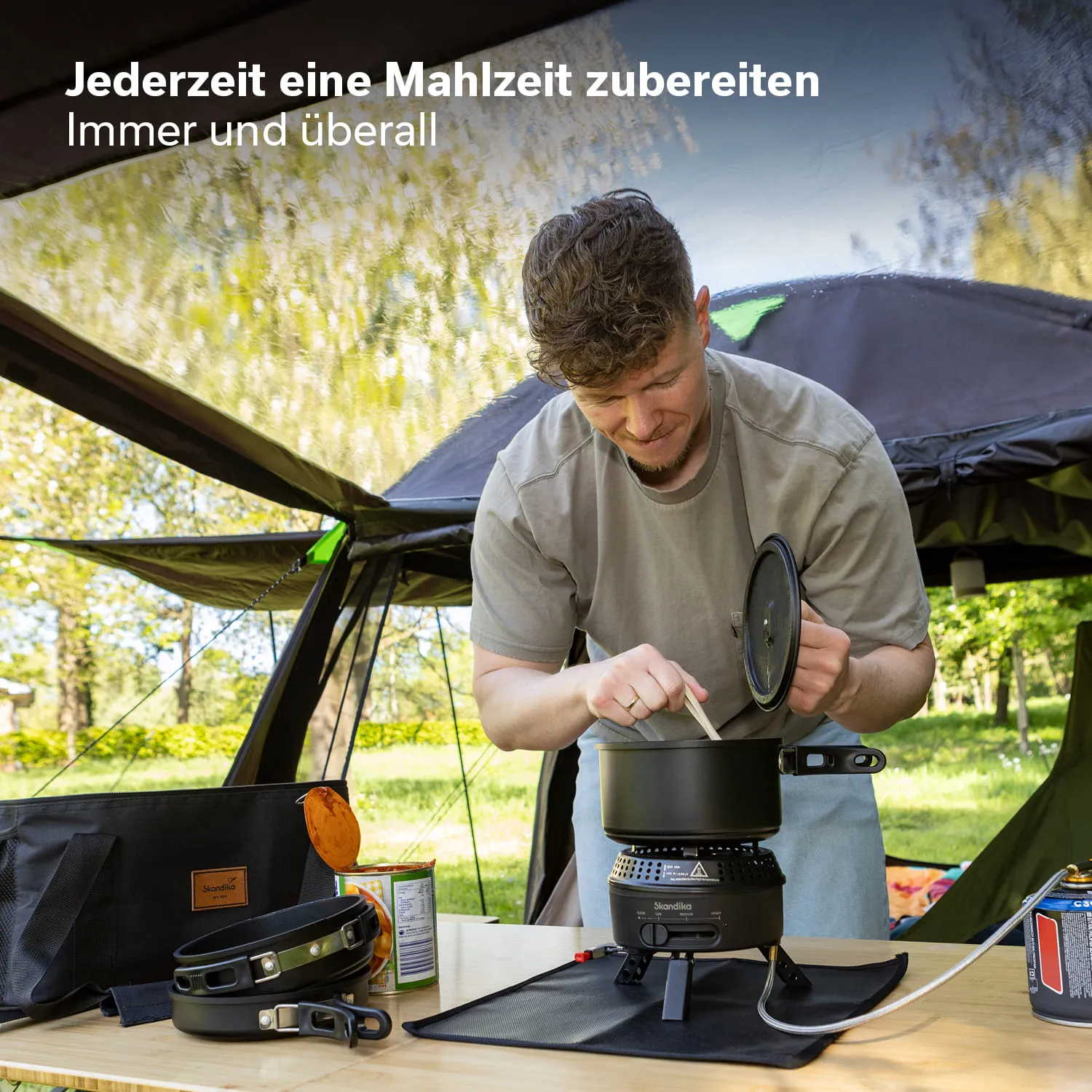 Person steht an einem Tisch im Freien und kocht mit einem Campingkocher. Neben ihm liegen Kochutensilien und eine Dose.