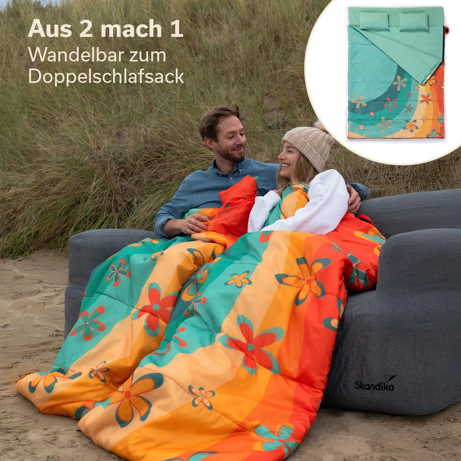Zwei Personen sitzen auf einem grauen Sofa am Strand, eingekuschelt in einen bunten Schlafsack mit Blumenmuster. Im Hintergrund sind grüne Gräser sichtbar. Oben rechts ist eine Darstellung eines aufgeschlagenen Schlafsacks.